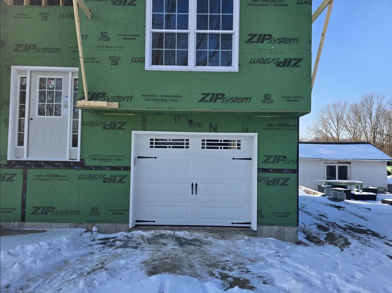 Front of a house under construction with green building wrap, white garage door, and a small front door with windows, snow on the ground, and another small building visible in the background.