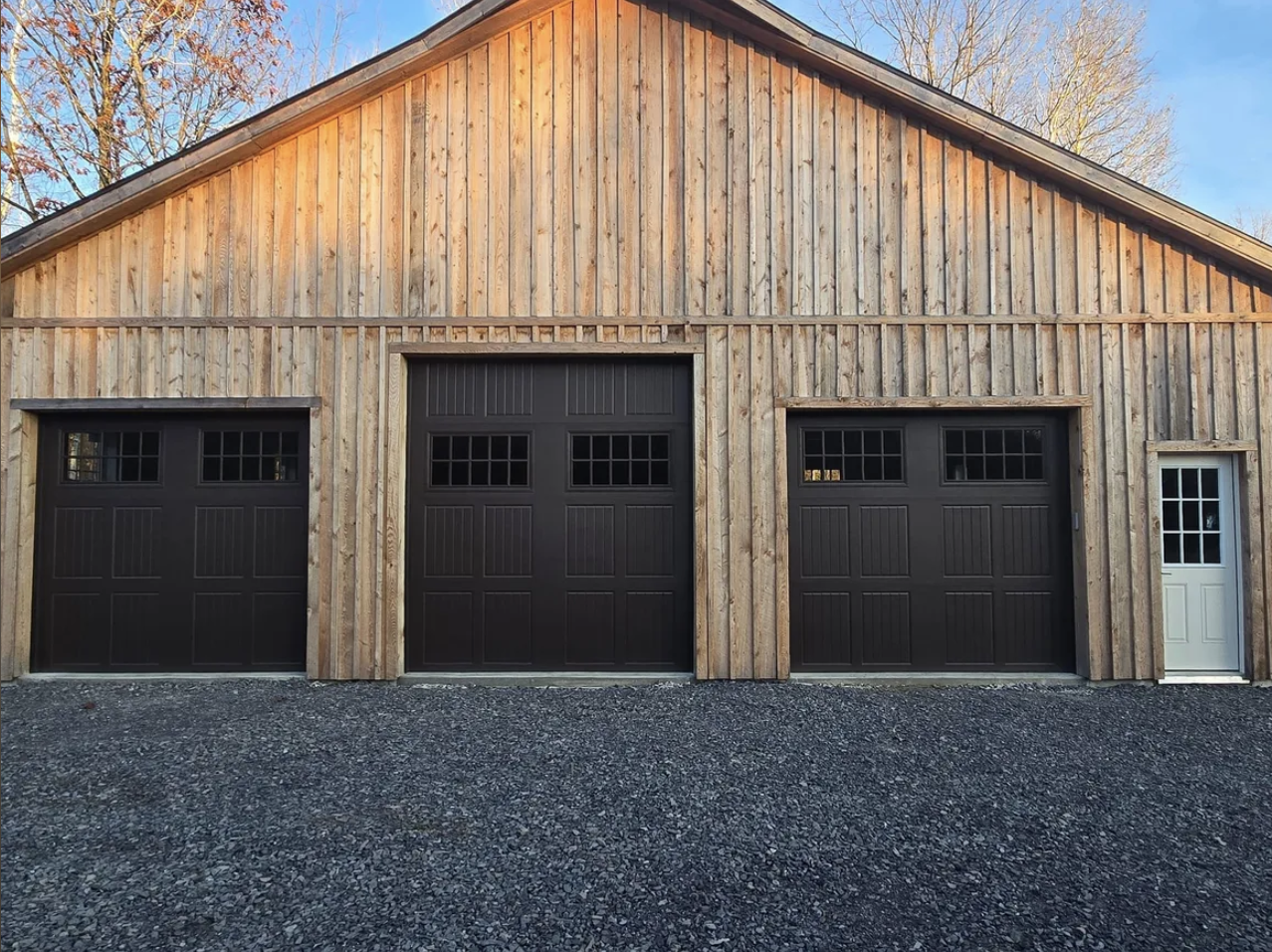 A large wooden garage with three doors: two black garage doors with small windows at the top and a white side door with glass panels on the right, all set against a gravel driveway and a background of trees.