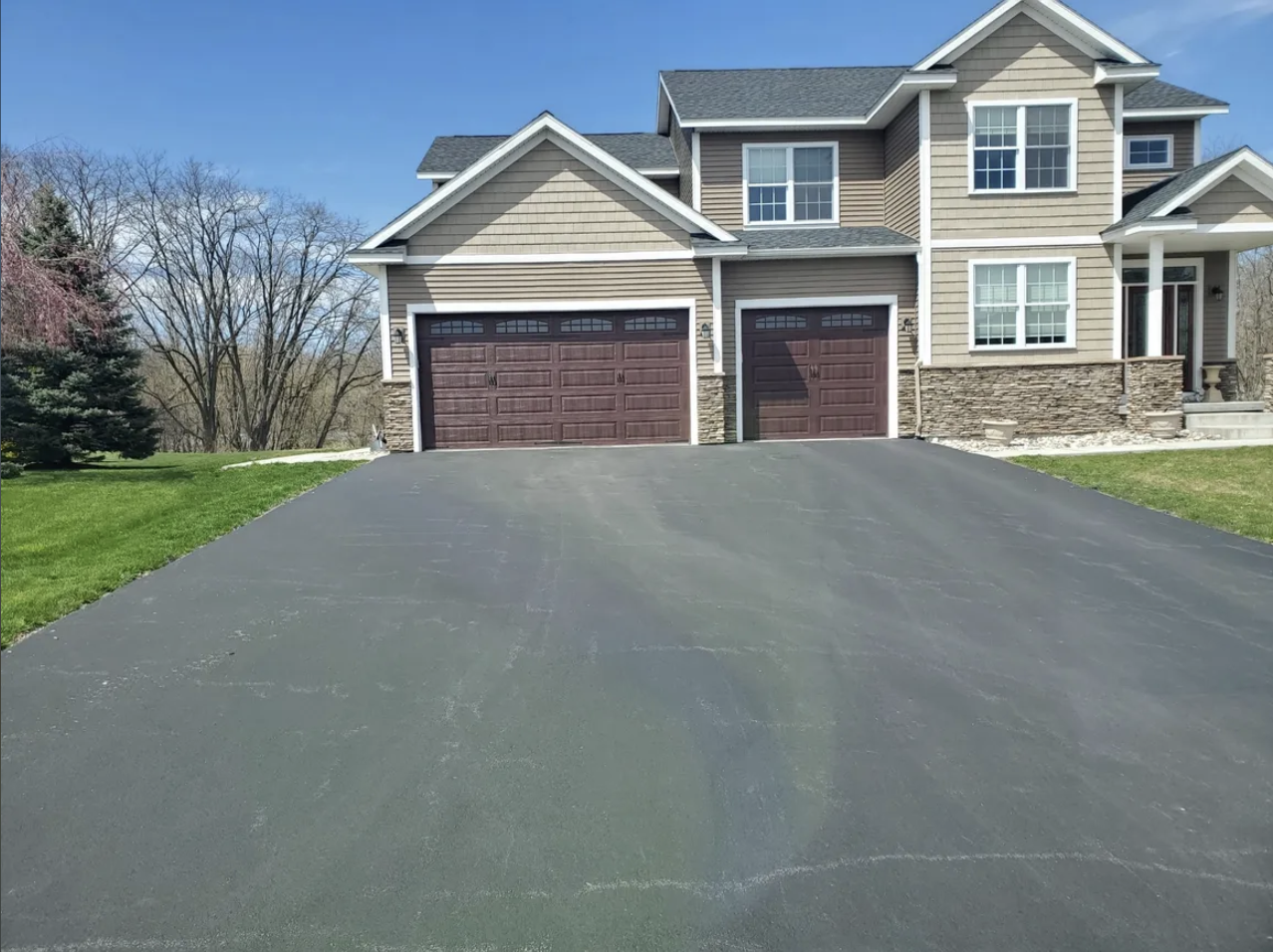A large two-story house with a double garage, beige siding with stone accents, white window frames, and a front porch, surrounded by a well-maintained lawn and trees, under a blue sky.