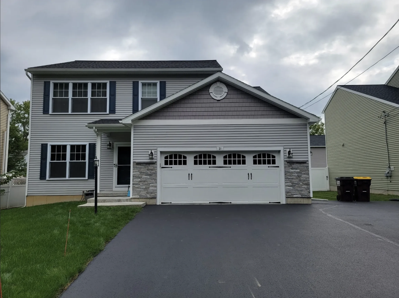 Front view of a two-story house with gray siding, dark shutters, and a double garage door. Grass lawn, driveway, and trash bins are visible.