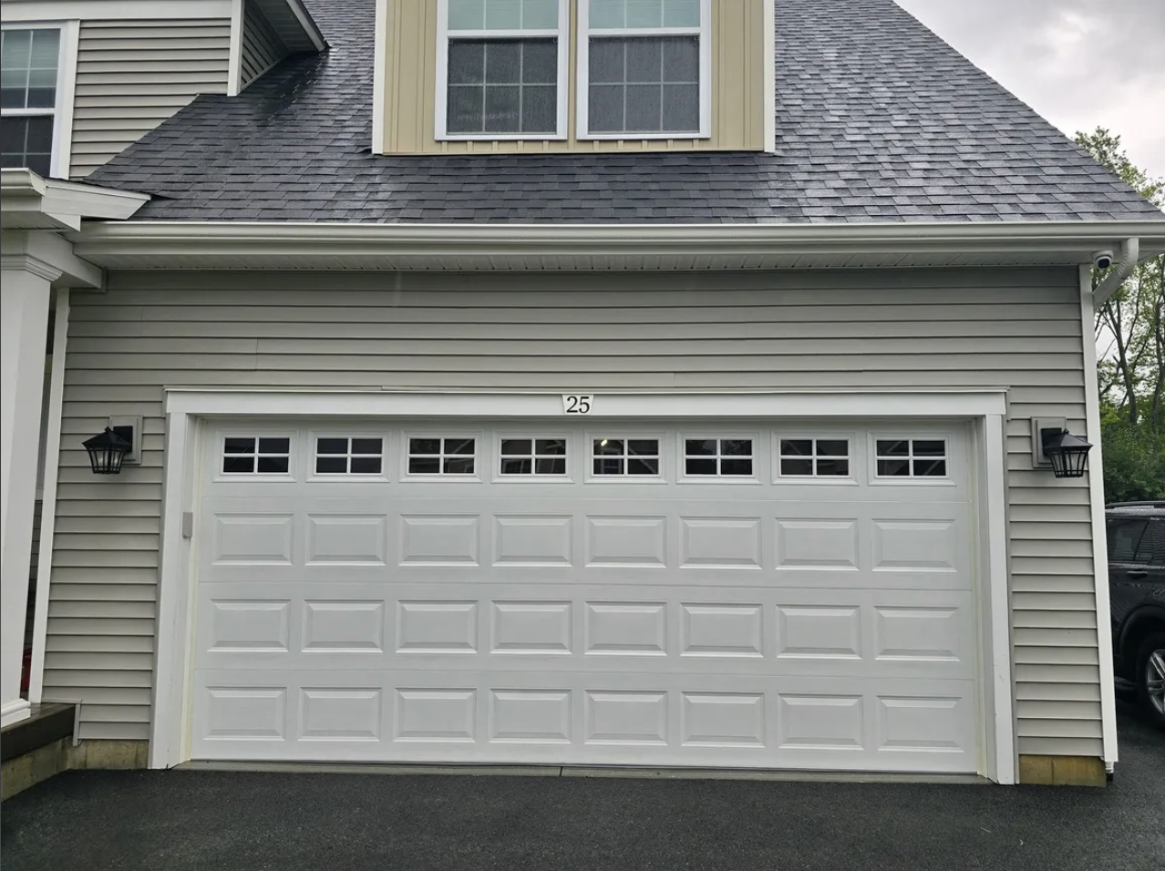 A white garage door with small rectangular windows at the top, framed by beige siding and an exterior wall with two black lantern-style light fixtures on either side, next to house number 25.