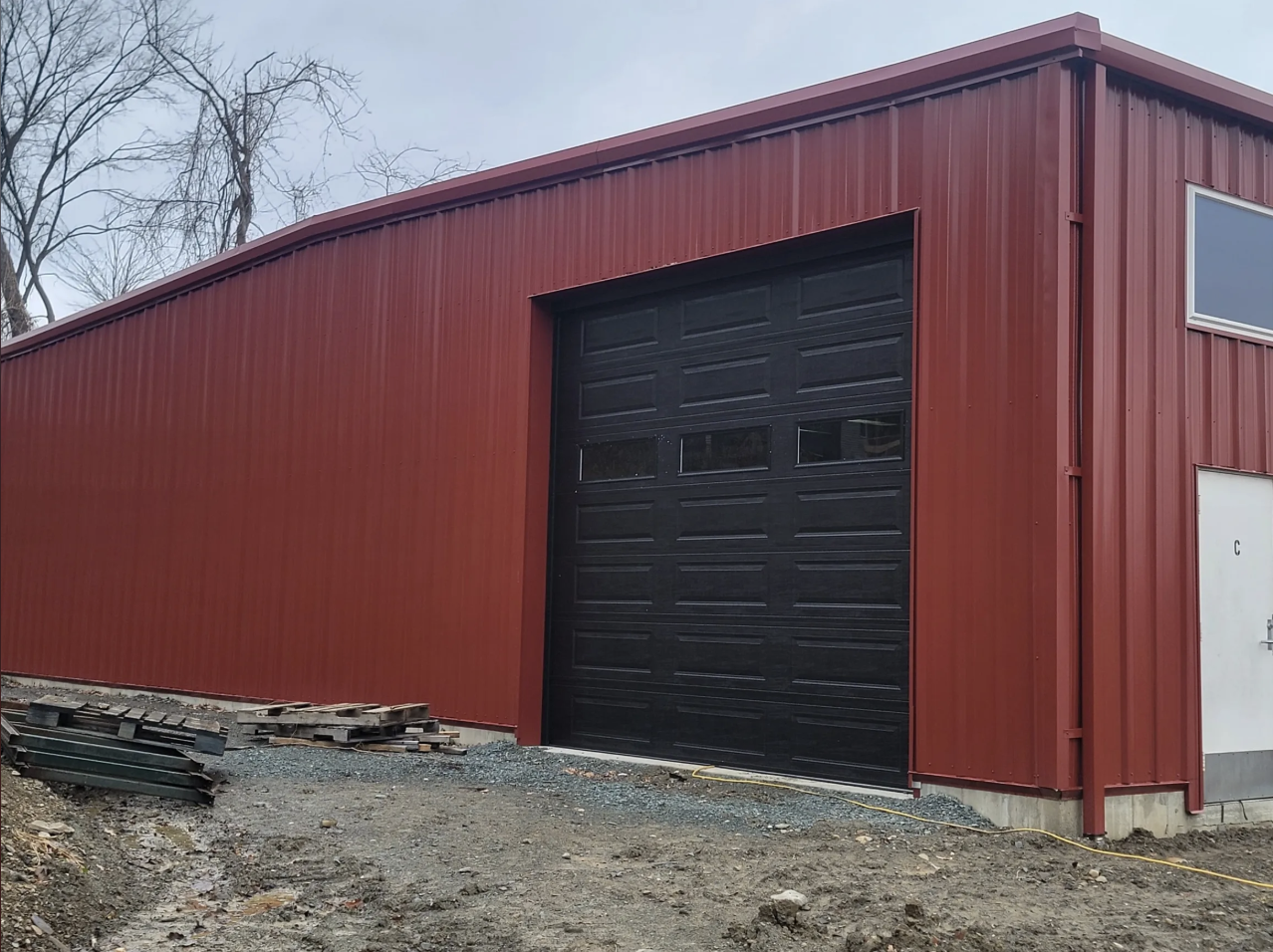 Red metal building with a large black garage door and a smaller white door, situated on uneven gravel ground with construction materials nearby, and leafless trees in the background under a cloudy sky.