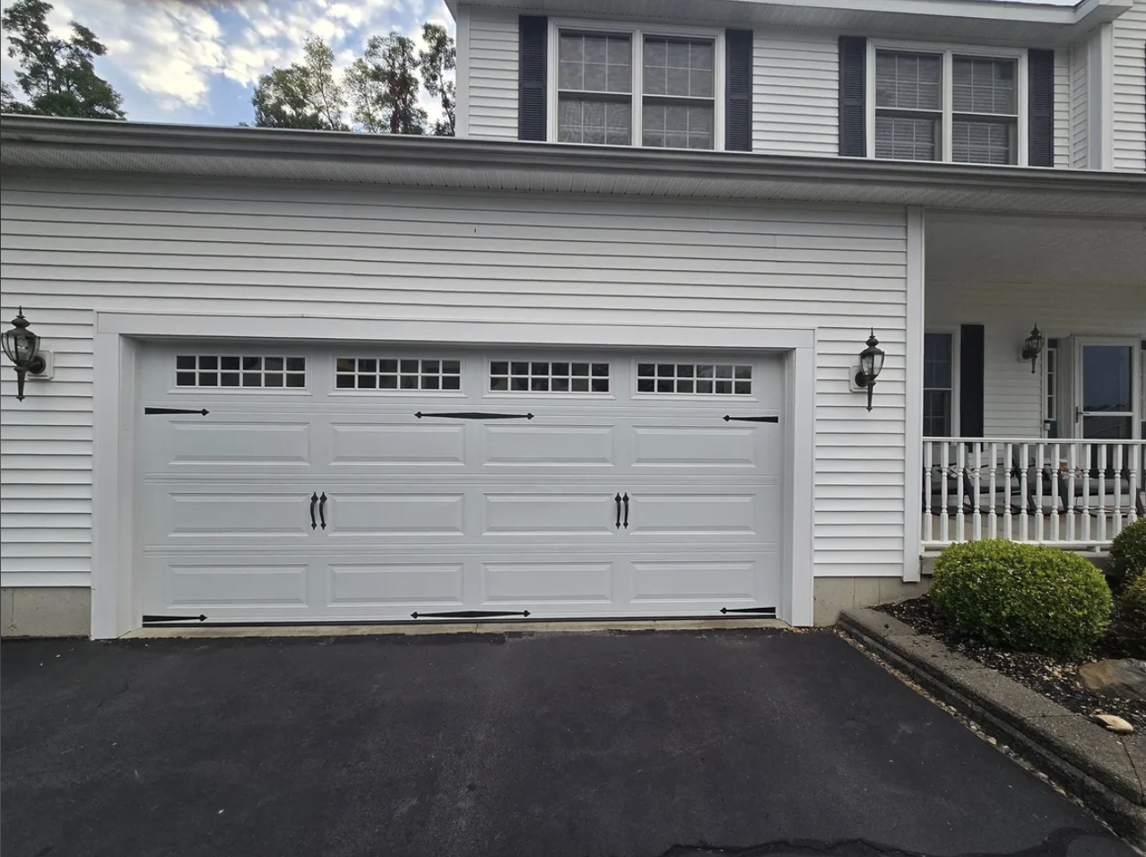 White garage door with windows at the top, black handles, and black decorative hinges, in front of a house with white siding, black shutters, and outdoor lanterns.