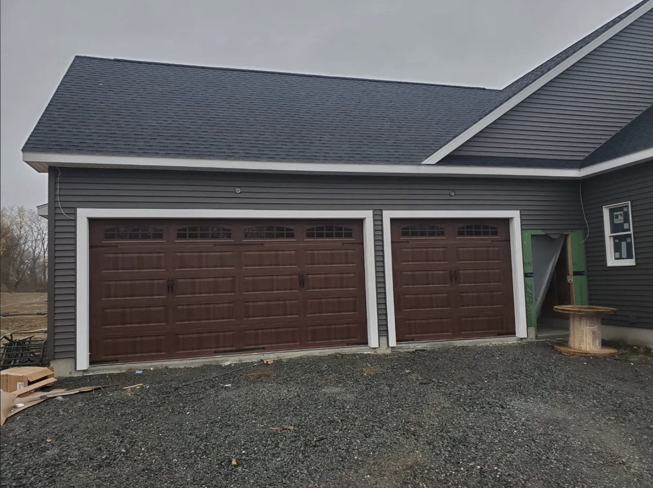 Newly constructed house with two brown garage doors, gray siding, and a gray shingle roof. There is a small window on the right side and construction materials and dirt in front.