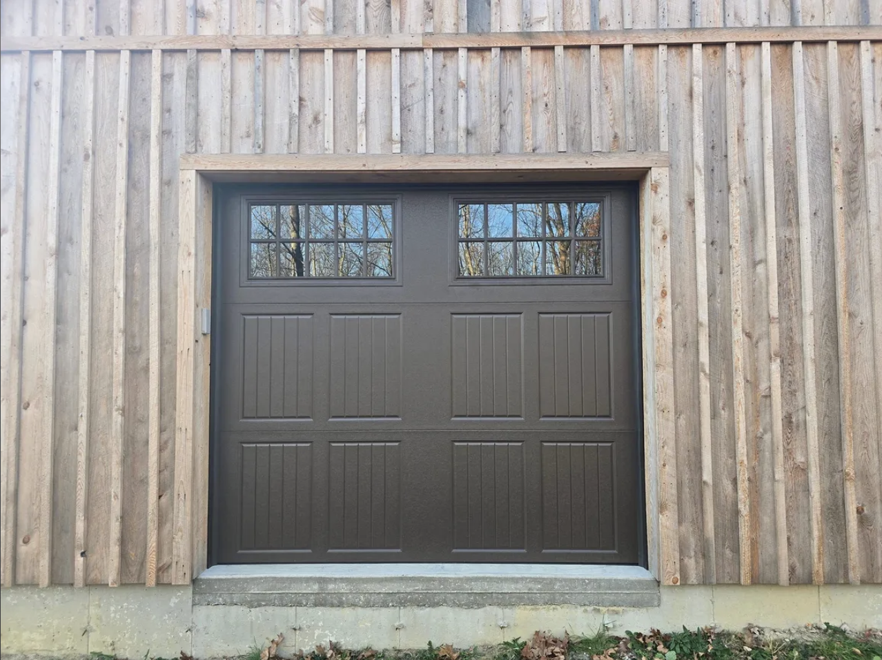 A dark gray garage door with rectangular signs of wood grain, set in a wooden building with vertical wood siding, and a concrete step in front.