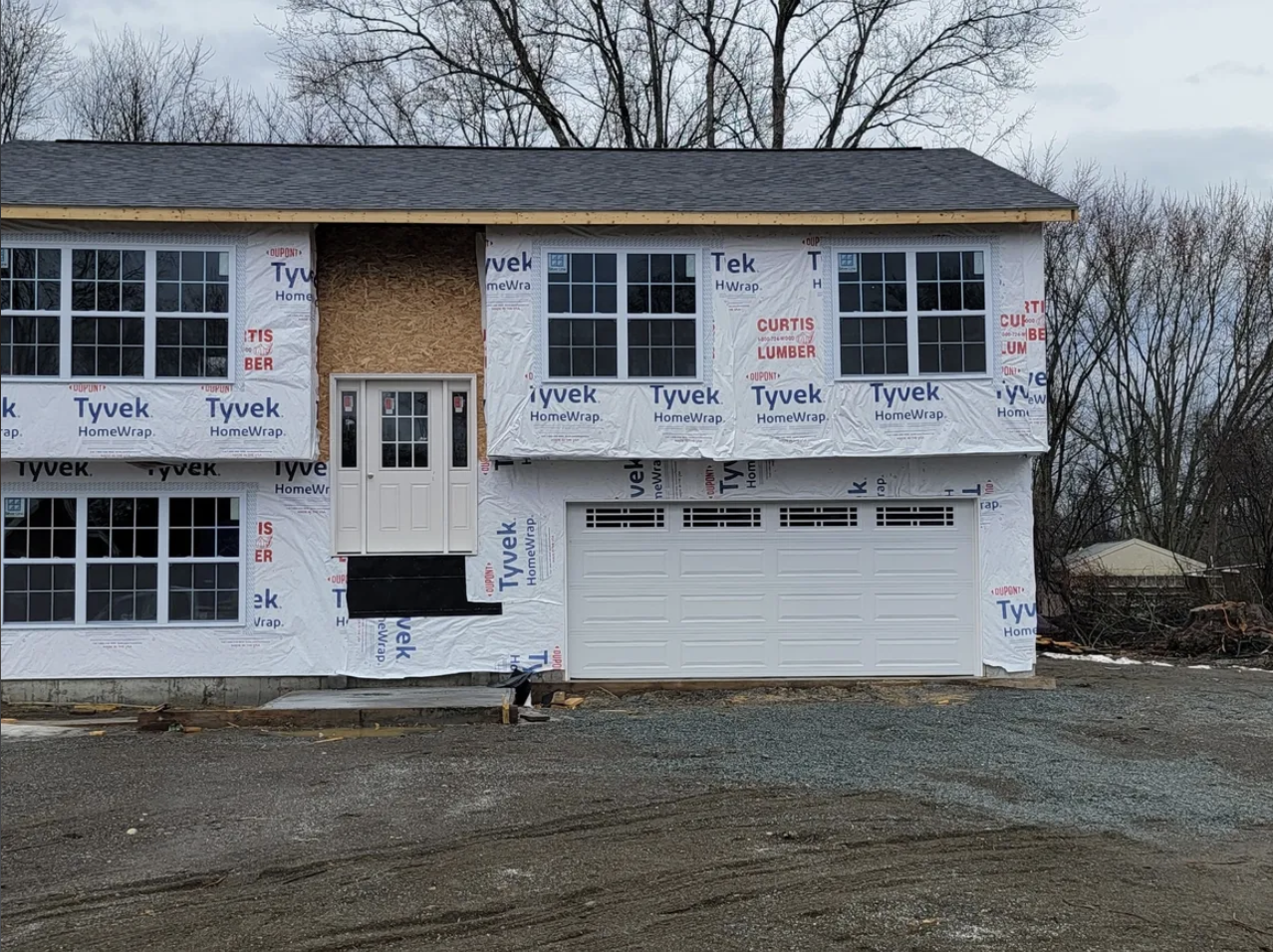 House under construction with siding partially installed, white garage door, front door with glass panels, multiple window openings, and construction materials on the ground.
