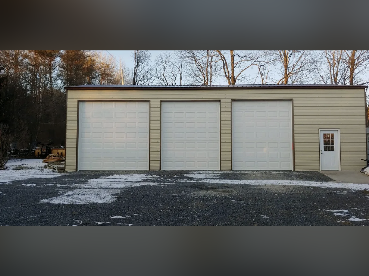 A beige metal building with three white garage doors and a small white side door, set against a backdrop of leafless trees and a partly cloudy sky, with some snow on the ground.