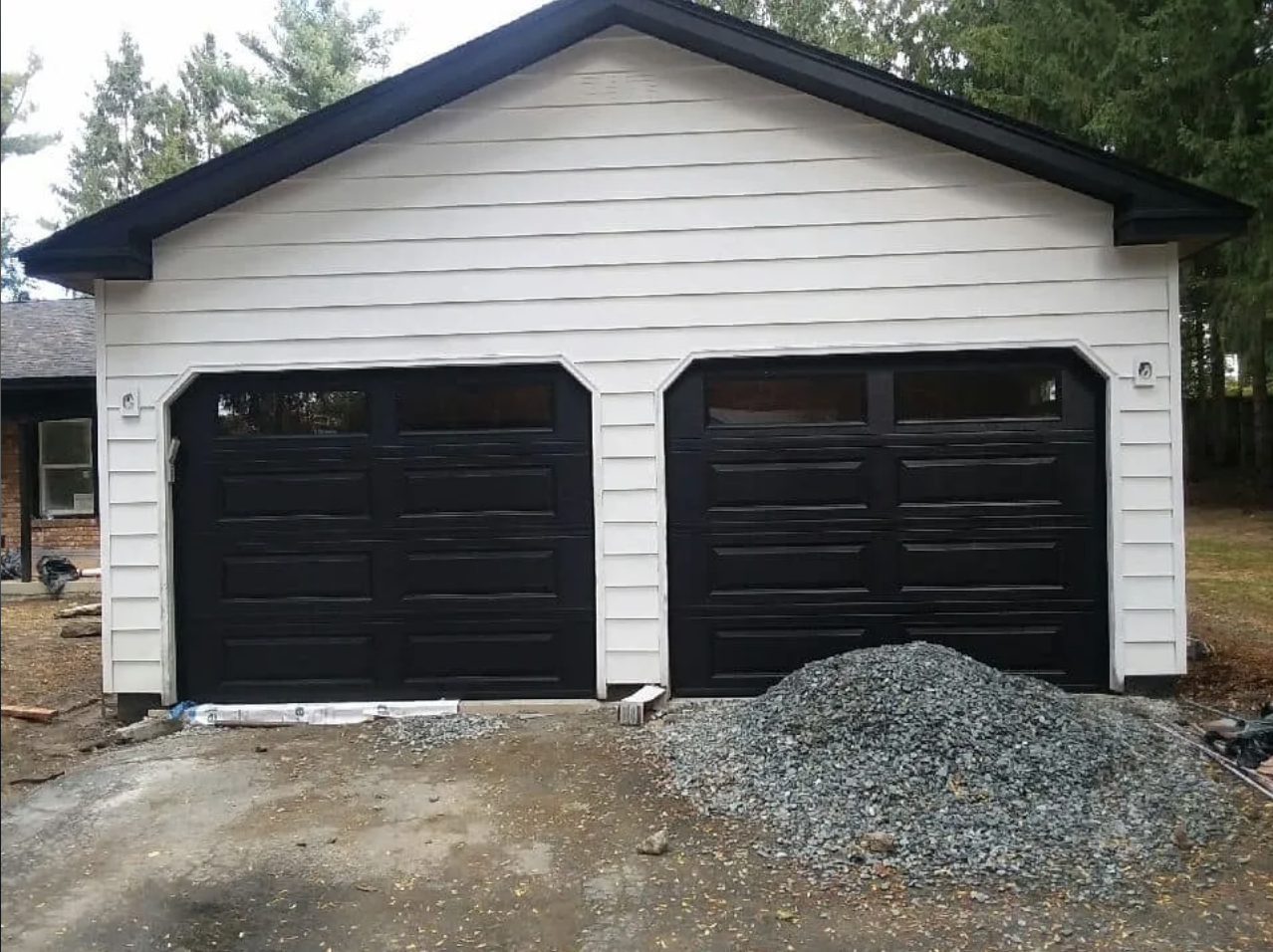 Garage with two closed black doors, white siding, and a pile of gravel in front, during daytime.