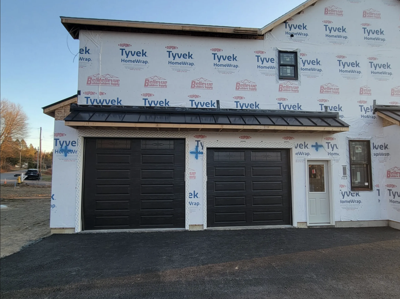 Newly constructed two-story house with vinyl siding in progress, featuring black garage doors, a small front door, and bay windows. Exterior wrapped with Tyvek HomeWrap for weatherproofing, with signs of ongoing work on the driveway and surrounding a