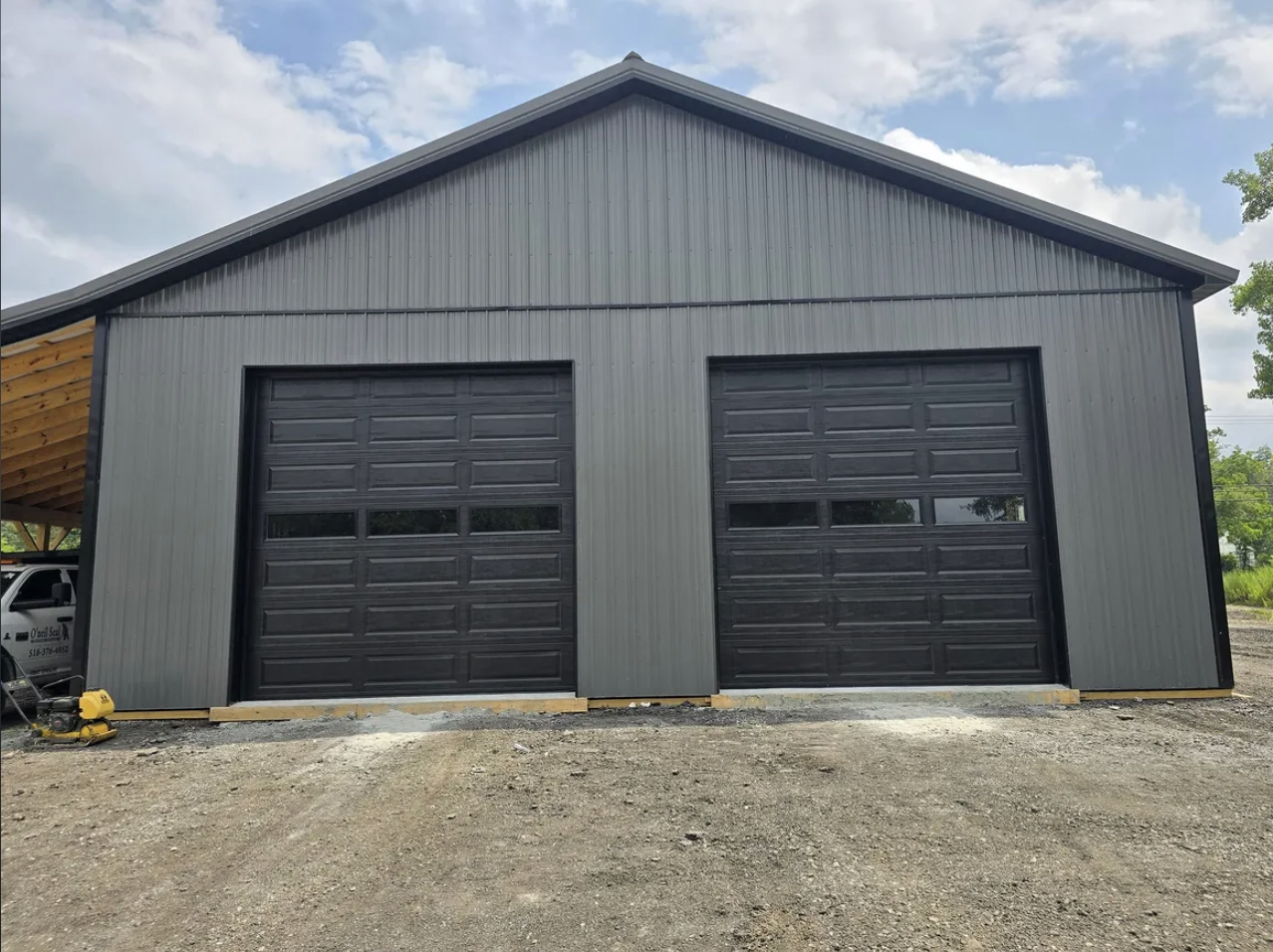 A large gray metal building with two black garage doors is under construction outdoors. The building is set on a dirt lot, with a partly cloudy sky overhead.