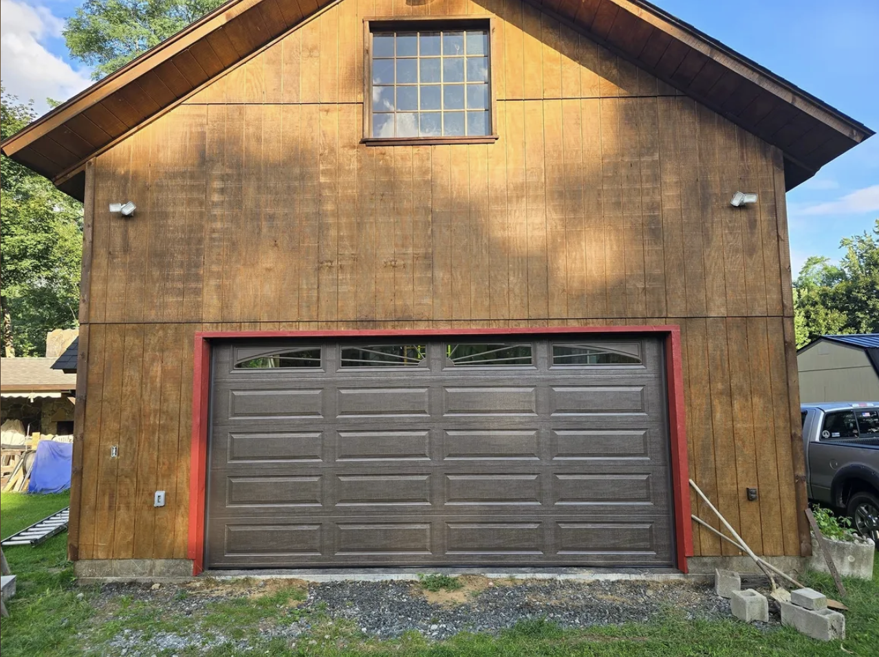 The front of a new garage with a closed garage door, wooden siding, and an upper window, surrounded by construction materials and greenery.