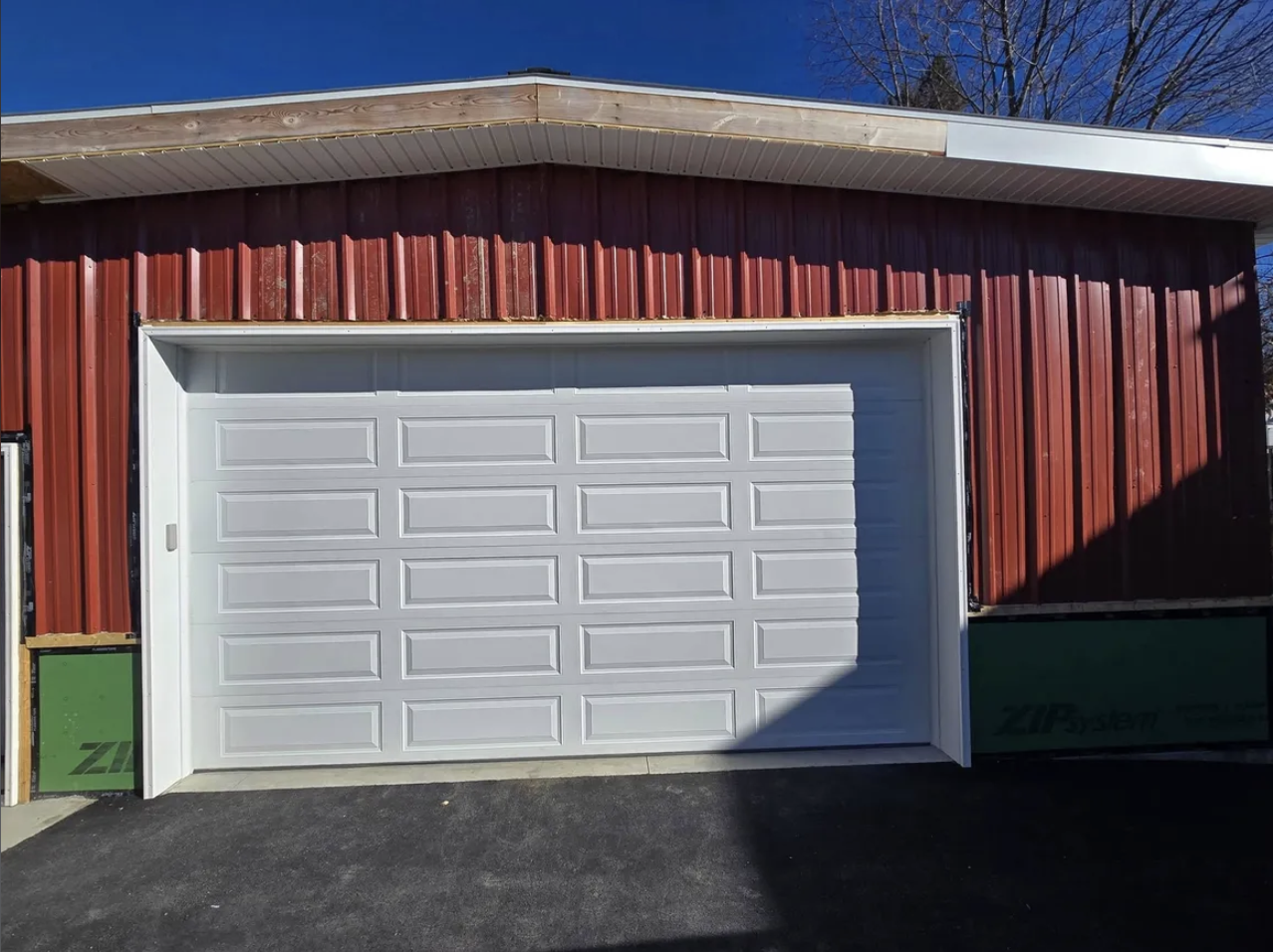 New white garage door on a red metal building, with a clear blue sky and shadows on the asphalt driveway.