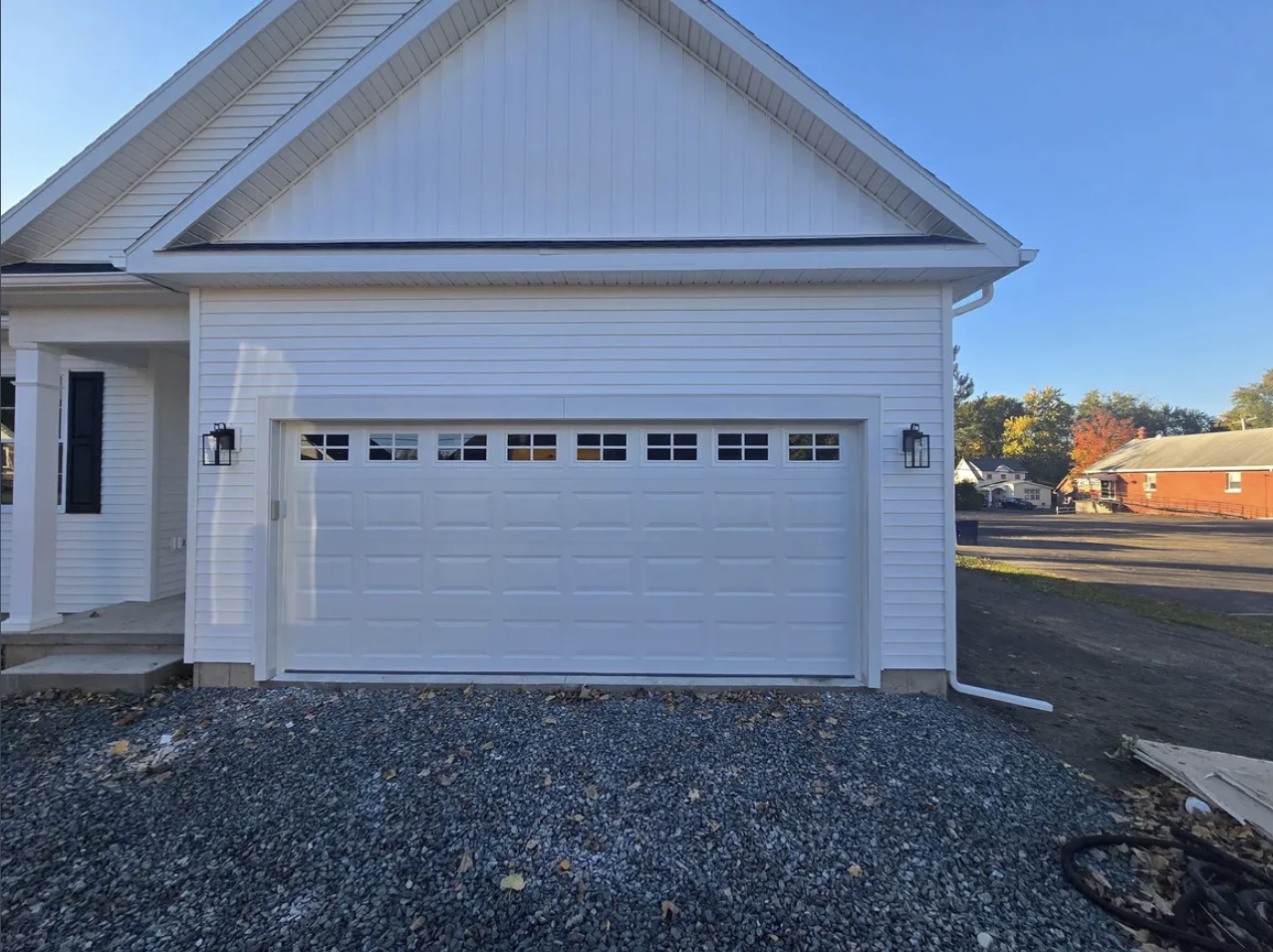 Front view of a white house with a closed garage door, black exterior lights, and a gravel driveway, with neighboring houses and trees in the background under a clear blue sky.