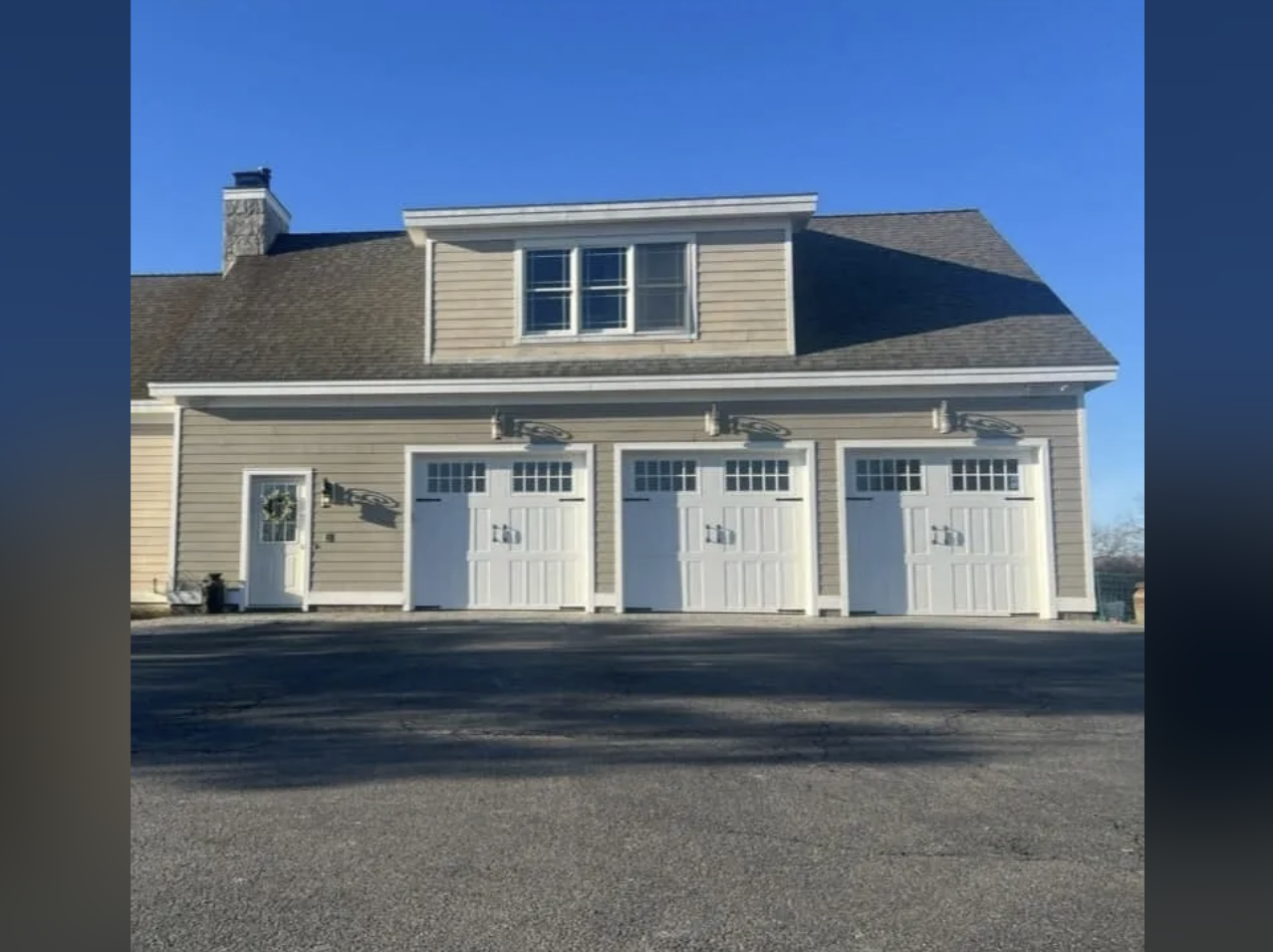 A multi-story house with three garage doors on the ground level, a small side door with a wreath, and a second story with three windows. The house has beige siding, a brown shingle roof, and is set against a clear blue sky.