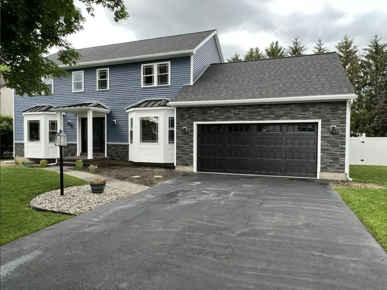 Front view of a modern two-story house with a black garage door, blue siding, and white accents. The driveway is asphalt, and there is a small landscaped area with a lamp post, and potted plants. The sky is overcast.