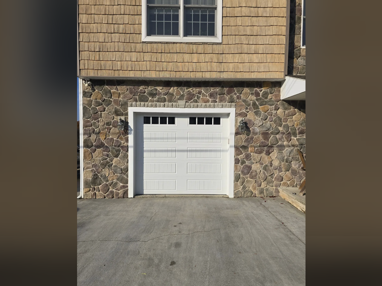 A white garage door set into a wall with stone veneer, flanked by two black outdoor wall lanterns, and a concrete driveway in front.