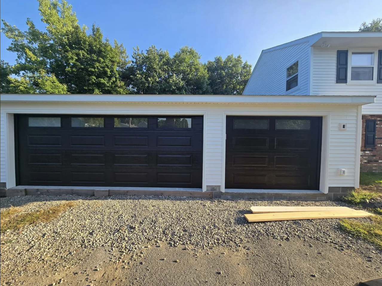 Front of a two-car garage with black doors, part of a white house, gravel driveway, and some wooden planks on the ground.