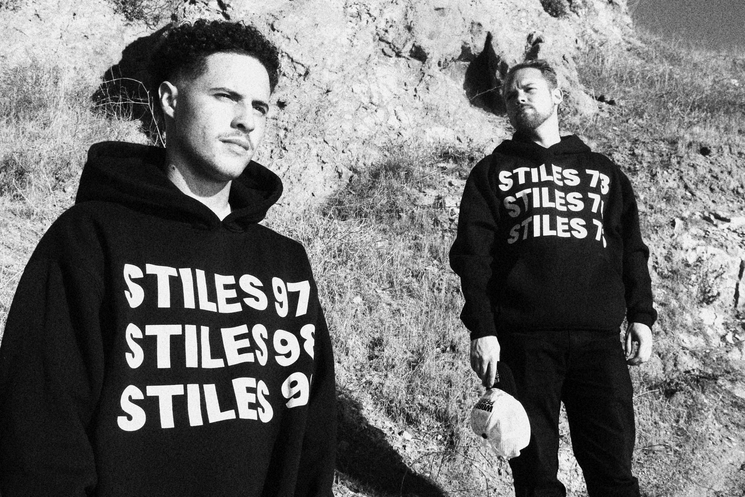 Two men standing outdoors in front of a rocky hillside, both wearing black hoodies with the text 'STILES 97' printed on them in large white letters. One man is holding a baseball cap in his right hand.