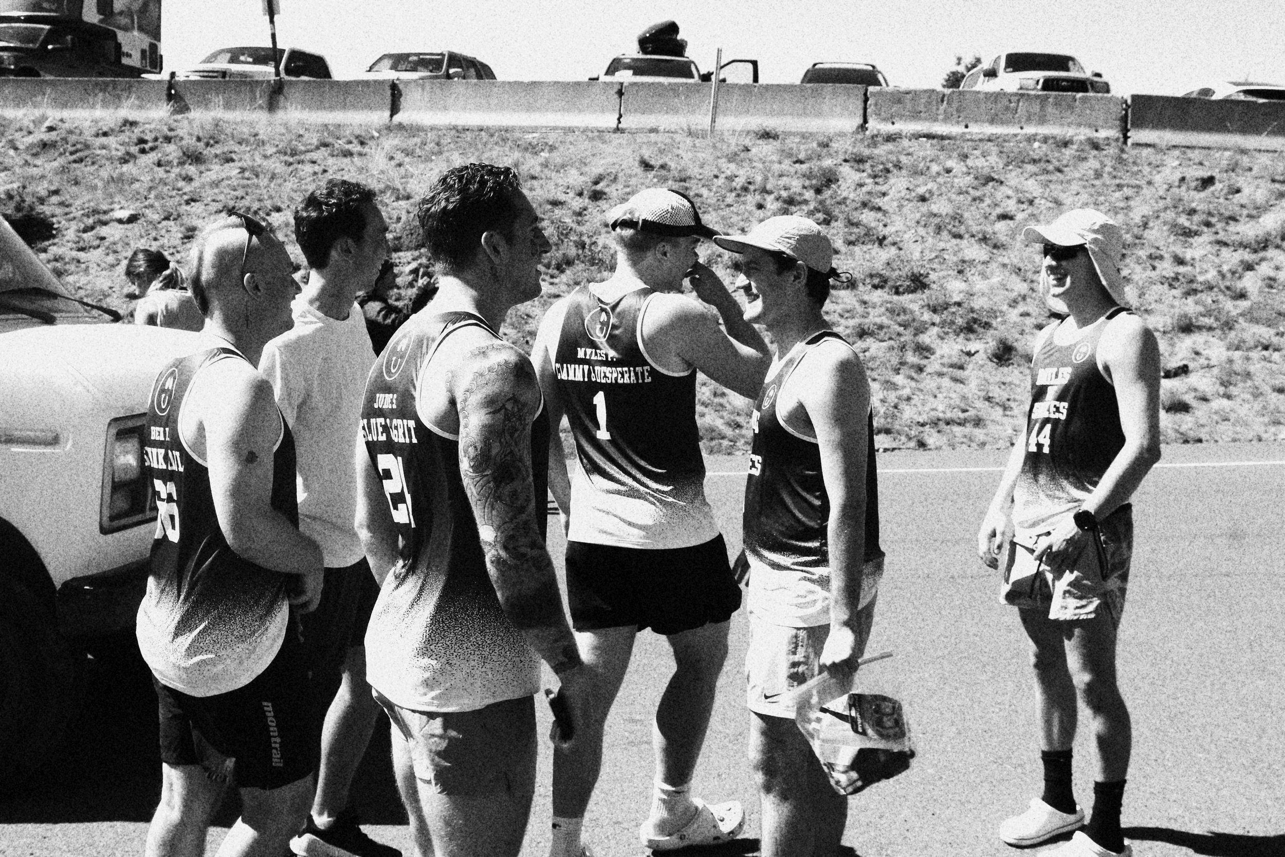 Group of seven people, six men and one woman, standing outdoors on a sunny day, engaged in conversation. Some are wearing athletic tank tops and shorts, with one holding a shoe. Background shows parked cars and a grassy hill.