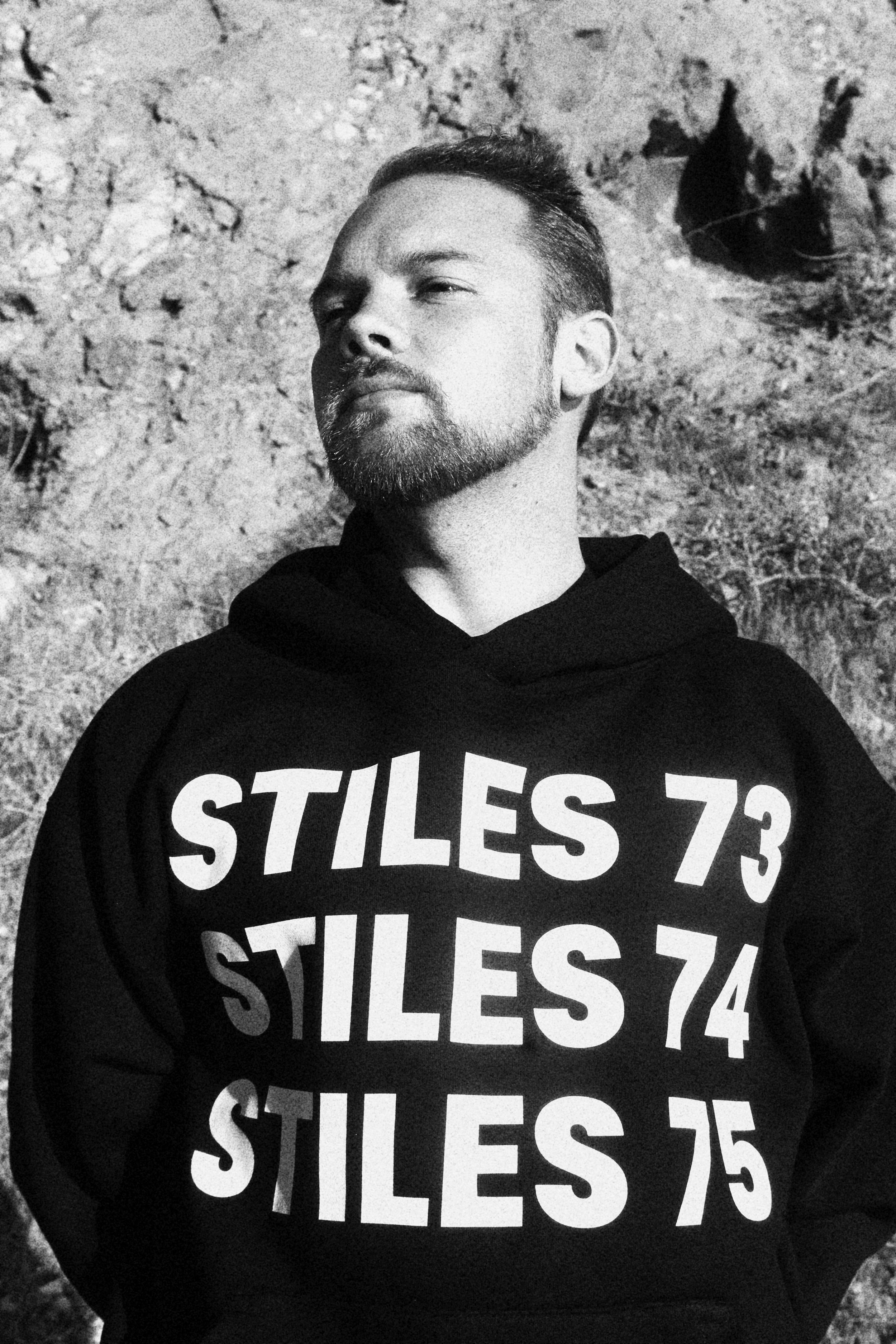 A black-and-white photo of a man with a beard and short hair, wearing a black hoodie with large white text that reads "STILES 73," "STILES 74," and "STILES 75" across the front. He is standing against a textured, natural background. 