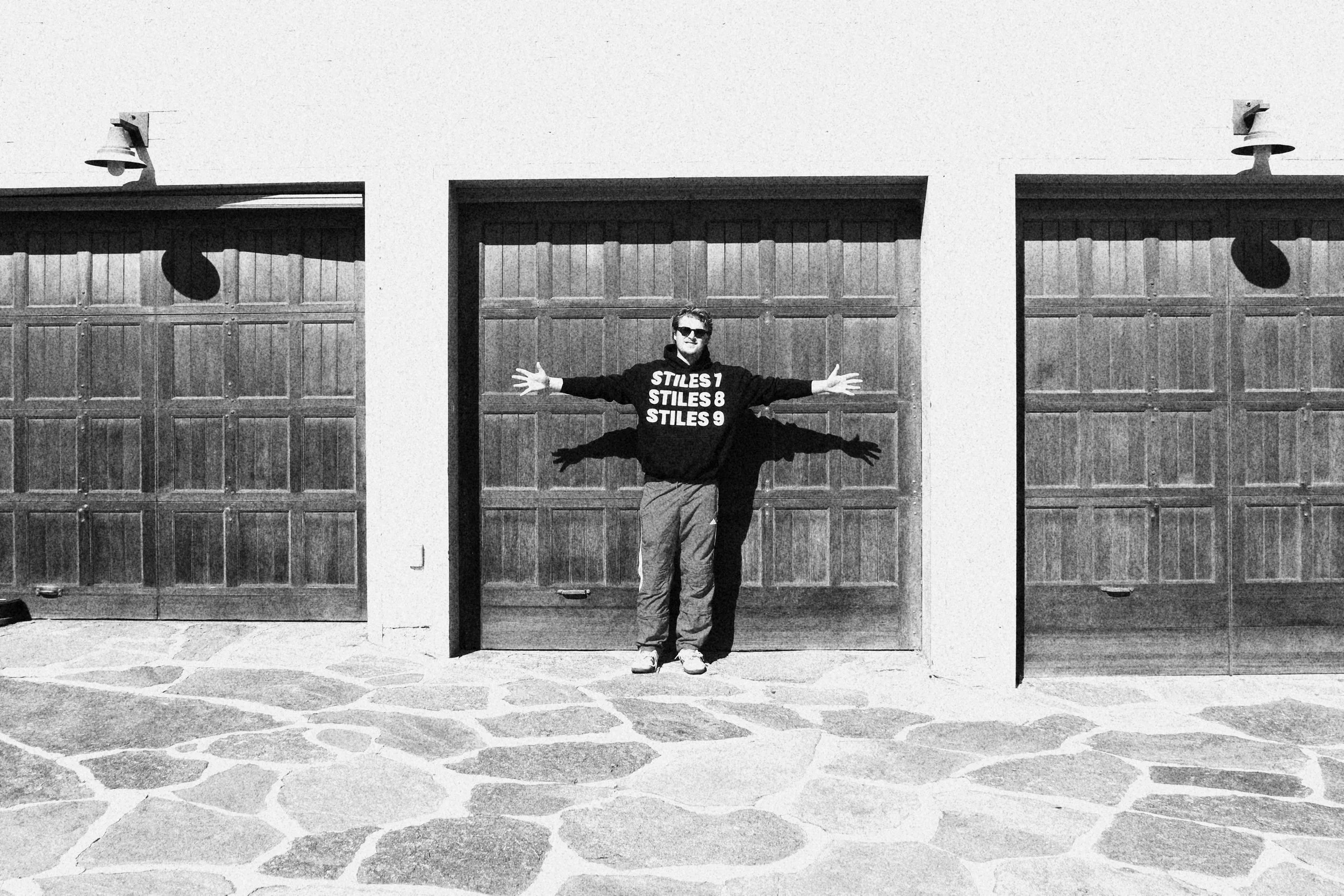 Person standing with outstretched arms in front of three wooden garage doors on a stone-paved ground, wearing sunglasses and a sweatshirt with text.