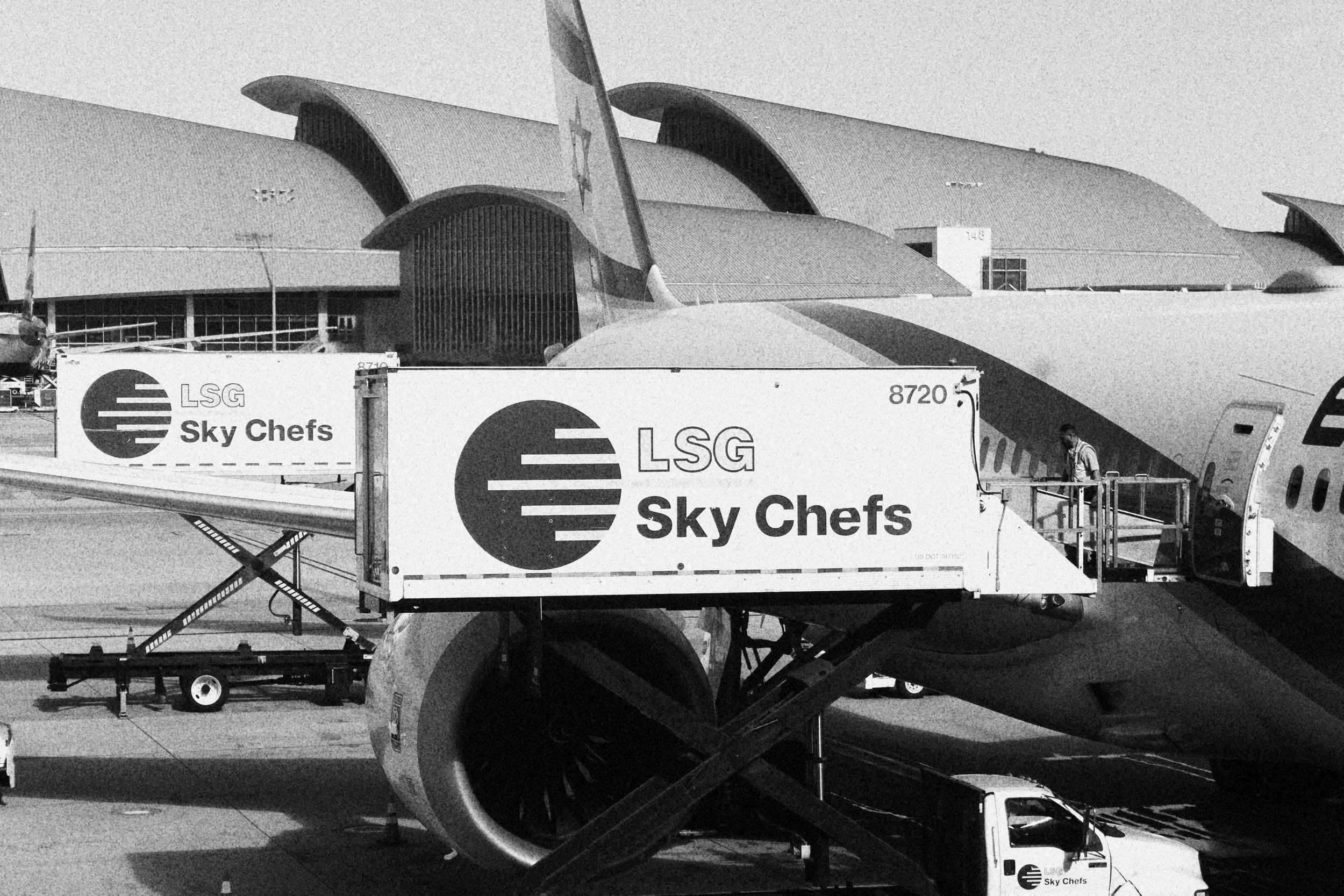 An airplane at an airport gate with Sky Chefs catering truck attached to its side.