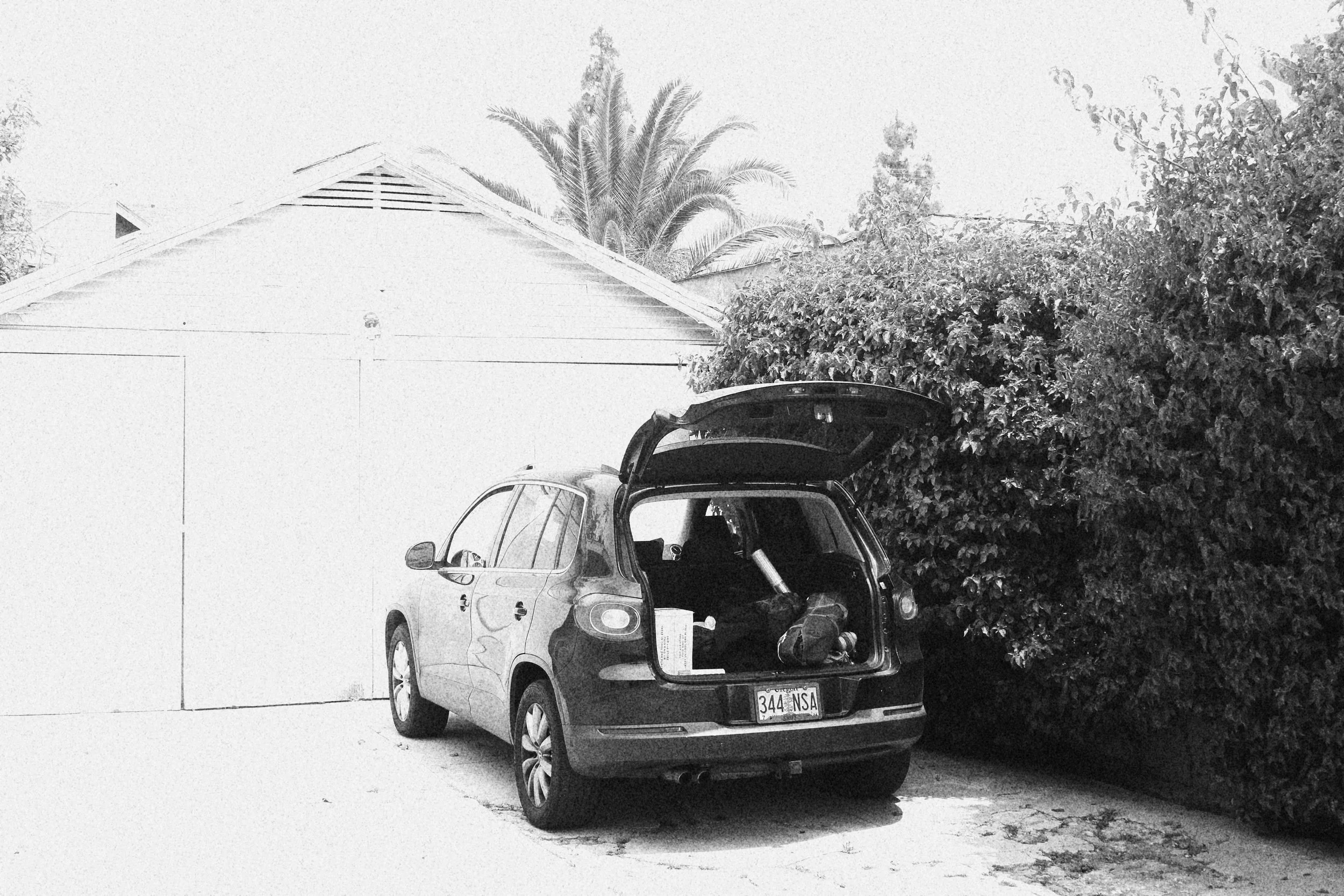 A car with its trunk open parked on a driveway next to dense bushes and a house with a gabled roof.