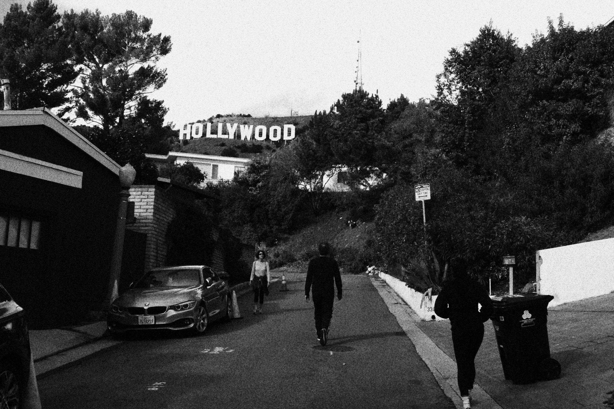 Black and white photo of a street view with the Hollywood sign on a hill in the background, trees and houses in the foreground, and three people walking along the street, one with a trash bin.