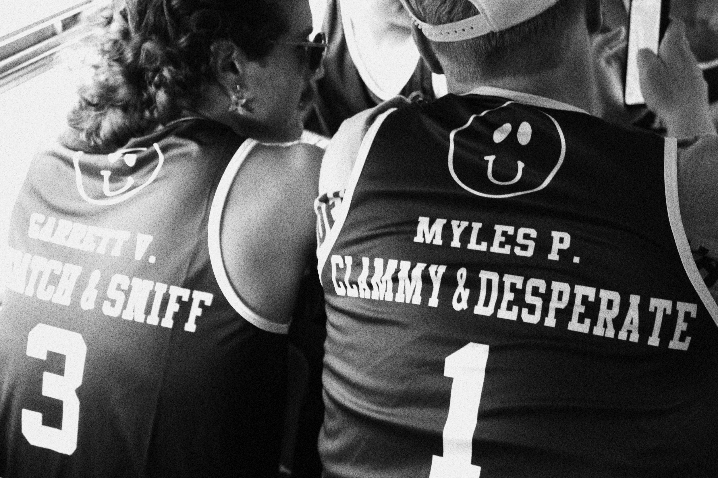 Back view of two women wearing sports jerseys with smiley face logos; the woman on the left has the name Charlotte and the number 3, the woman on the right has the name Myles P. and the number 1. They are close together, possibly in a team setting.
