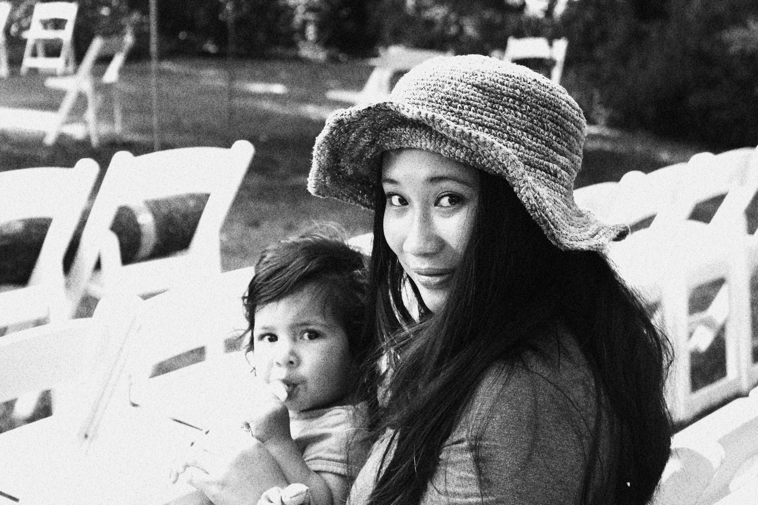 A woman with long dark hair wearing a large, textured hat, holding a young girl with short hair and a curious expression, in an outdoor setting with white chairs in the background.