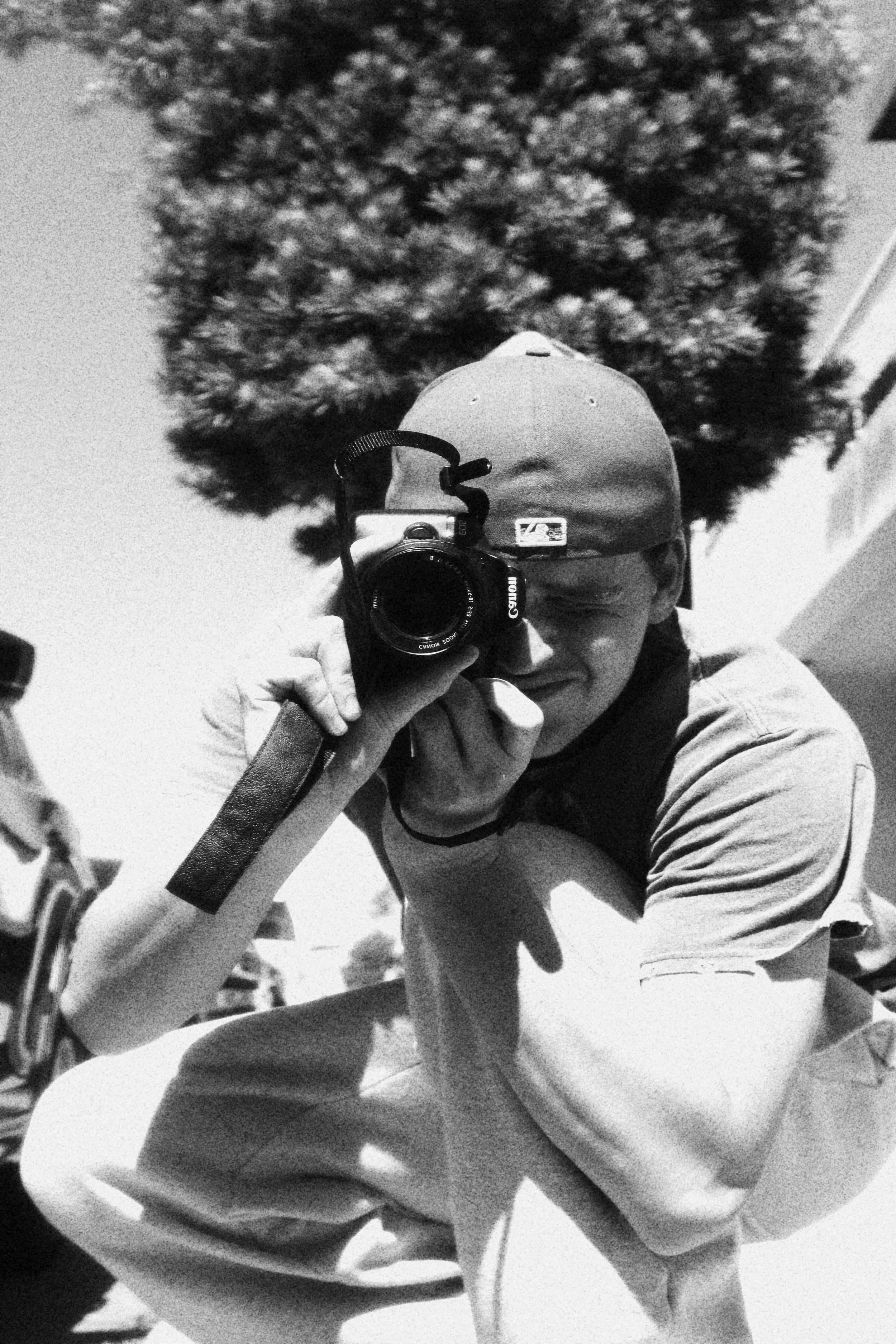 A young person taking a self-portrait with a camera in front of a mirror outdoors on a sunny day, with a large tree in the background.
