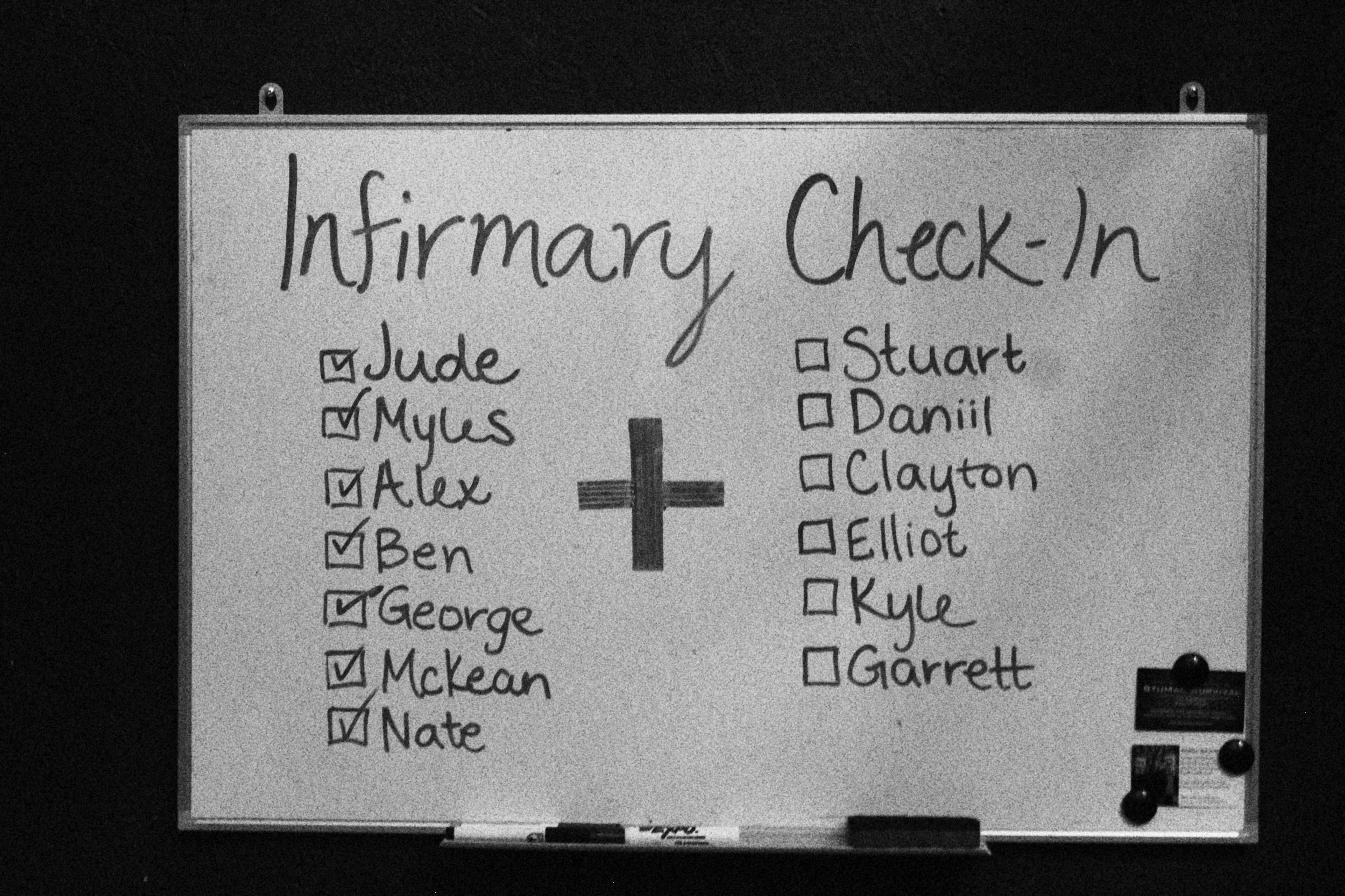 Whiteboard with a sign reading 'Infirmary Check-In' and a list of names, some checked off, divided into two columns by a plus sign, with a black background.