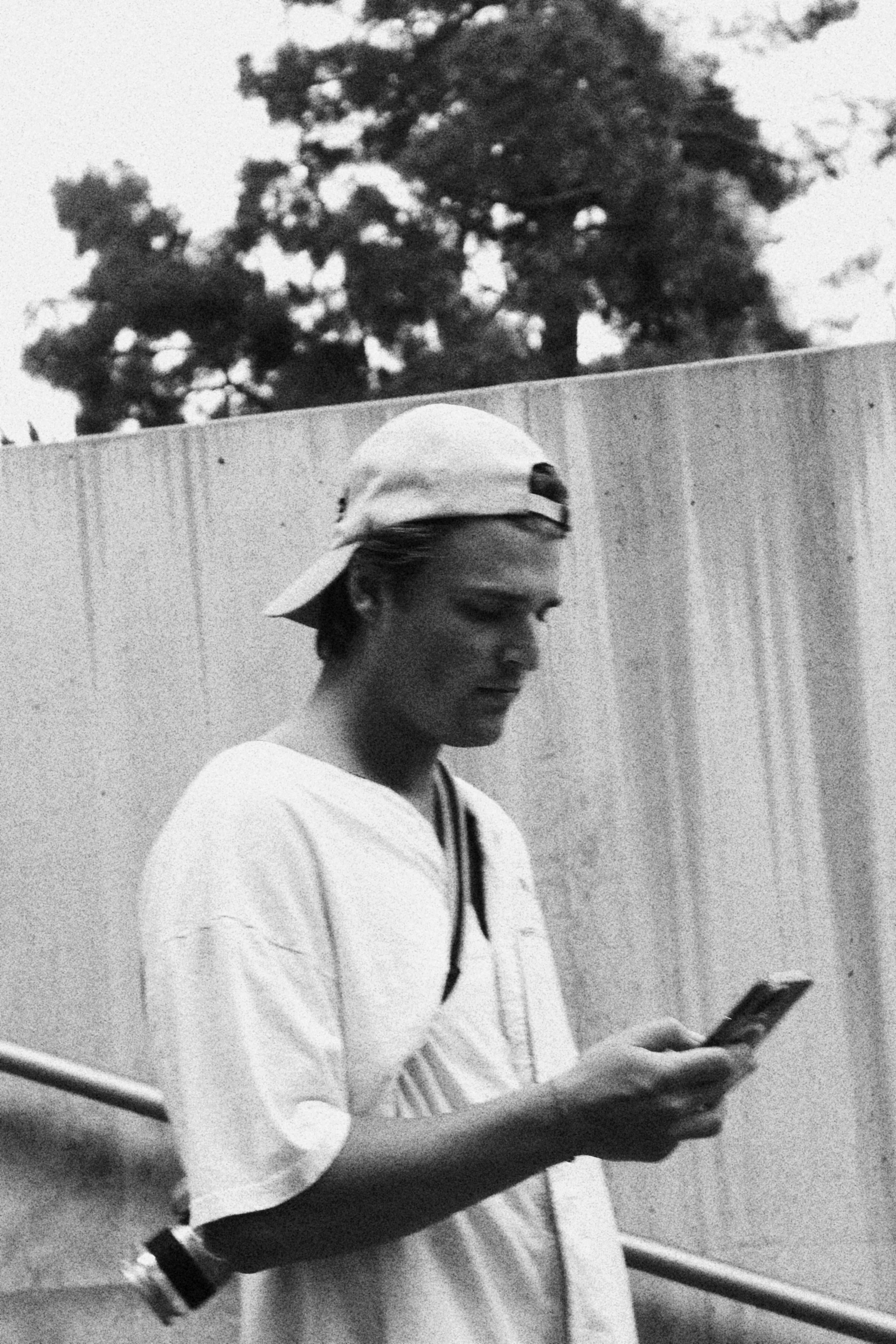 A young woman wearing a baseball cap backwards, a white t-shirt, and a wristwatch, looking at her phone outdoors near a wooden fence with trees in the background.