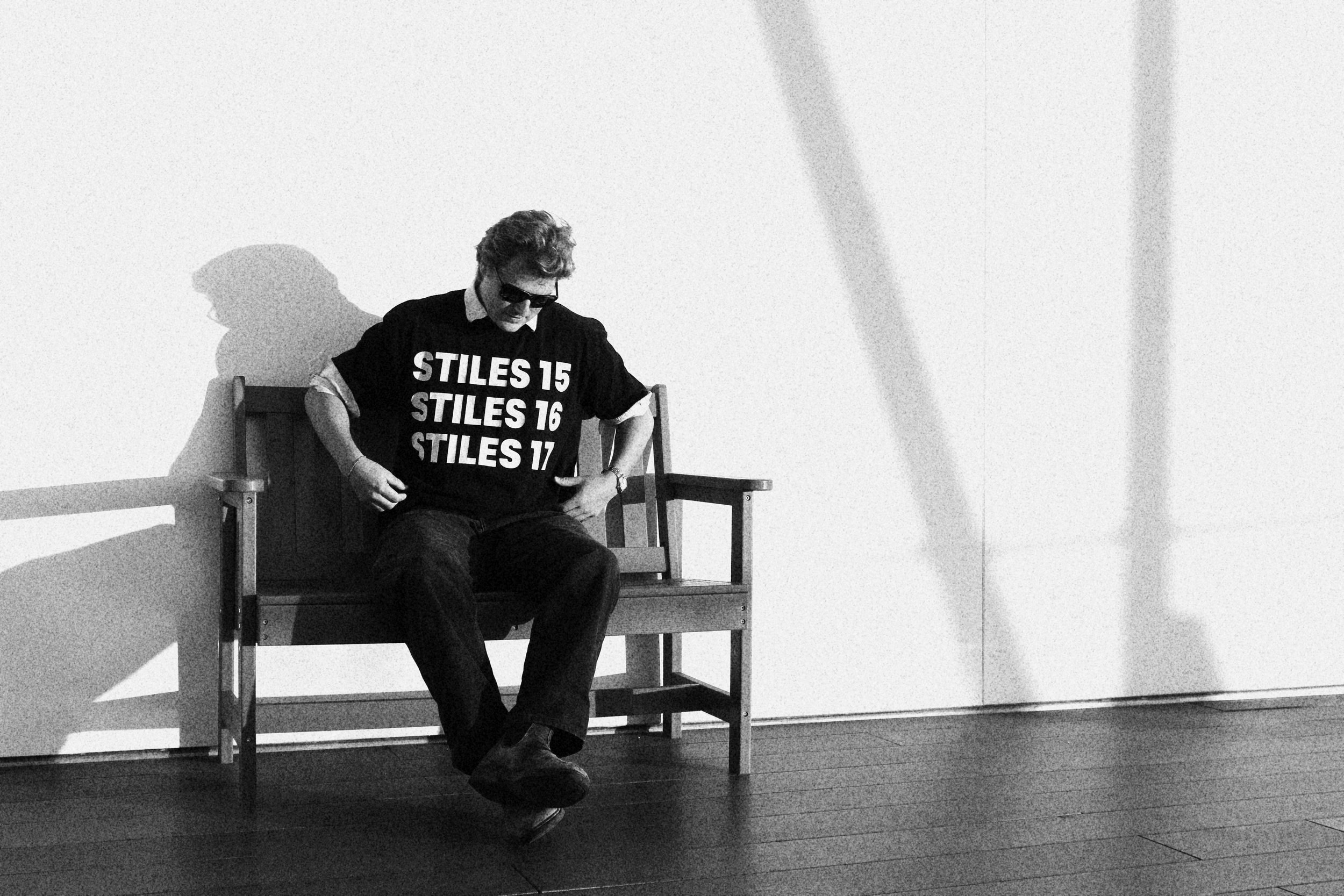 A man in sunglasses sitting on a wooden bench against a plain wall, wearing a black T-shirt with white text and dark pants. The man's head is tilted downward, and his hands rest on his thighs. His shadow is cast on the wall behind him.