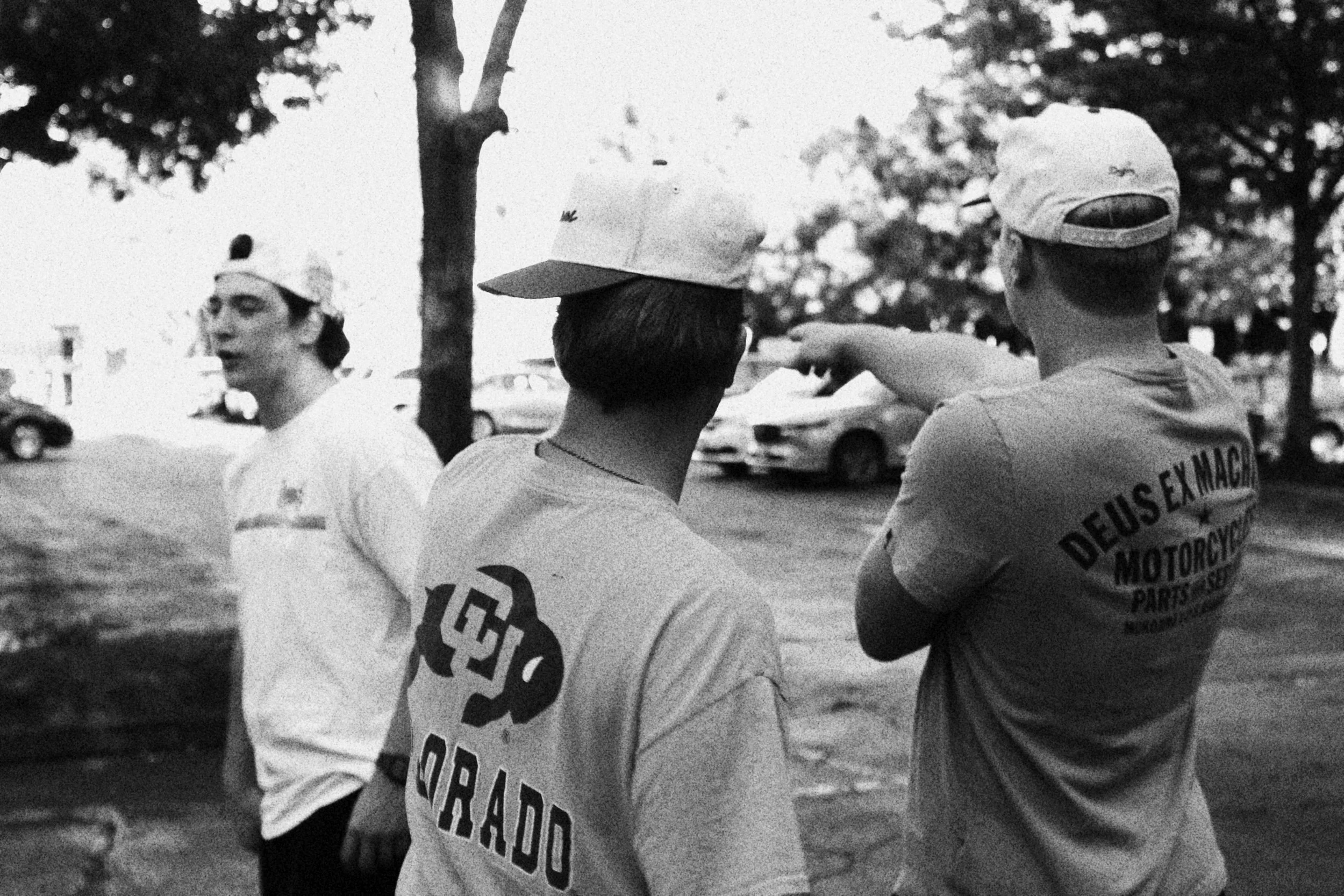 Three young men wearing caps and casual clothes stand outdoors near parked cars underneath trees, one of them is pointing.