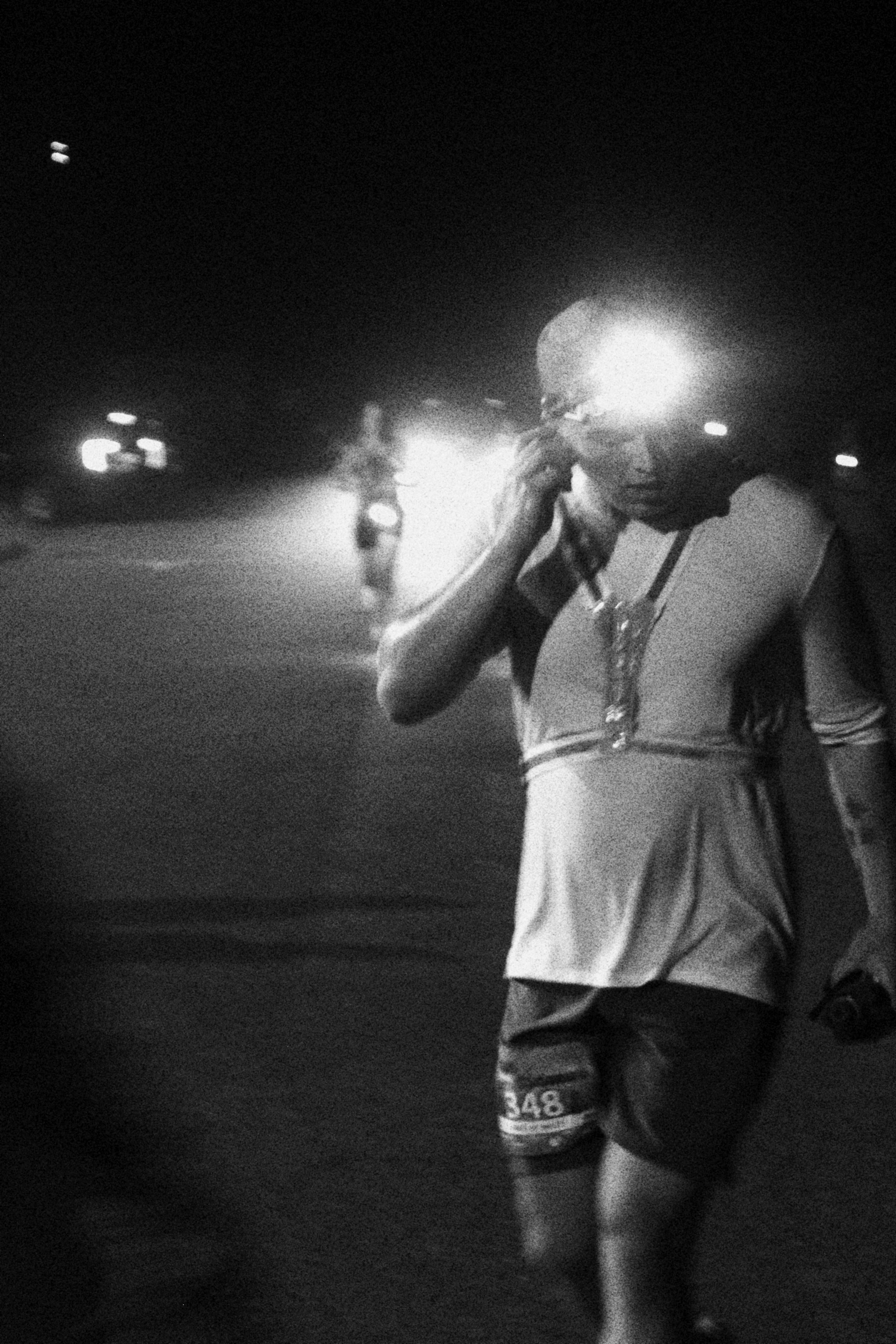 A person walking in a dark outdoor setting, wearing a race bib with the number 348, and holding an object in one hand, with bright lights and another person in the background.