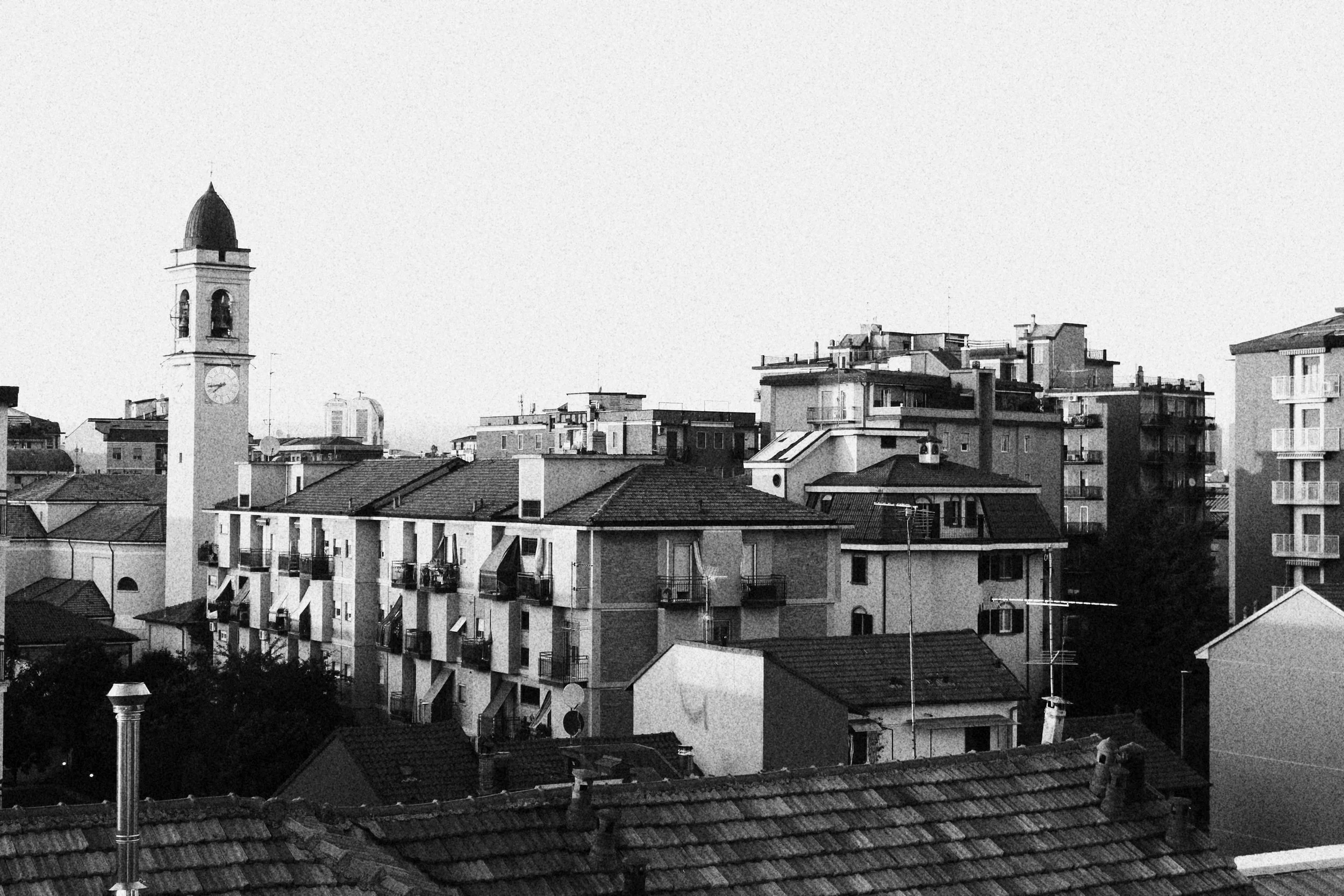 Black and white photo of a cityscape with apartment buildings, rooftops, and a clock tower with a spire.