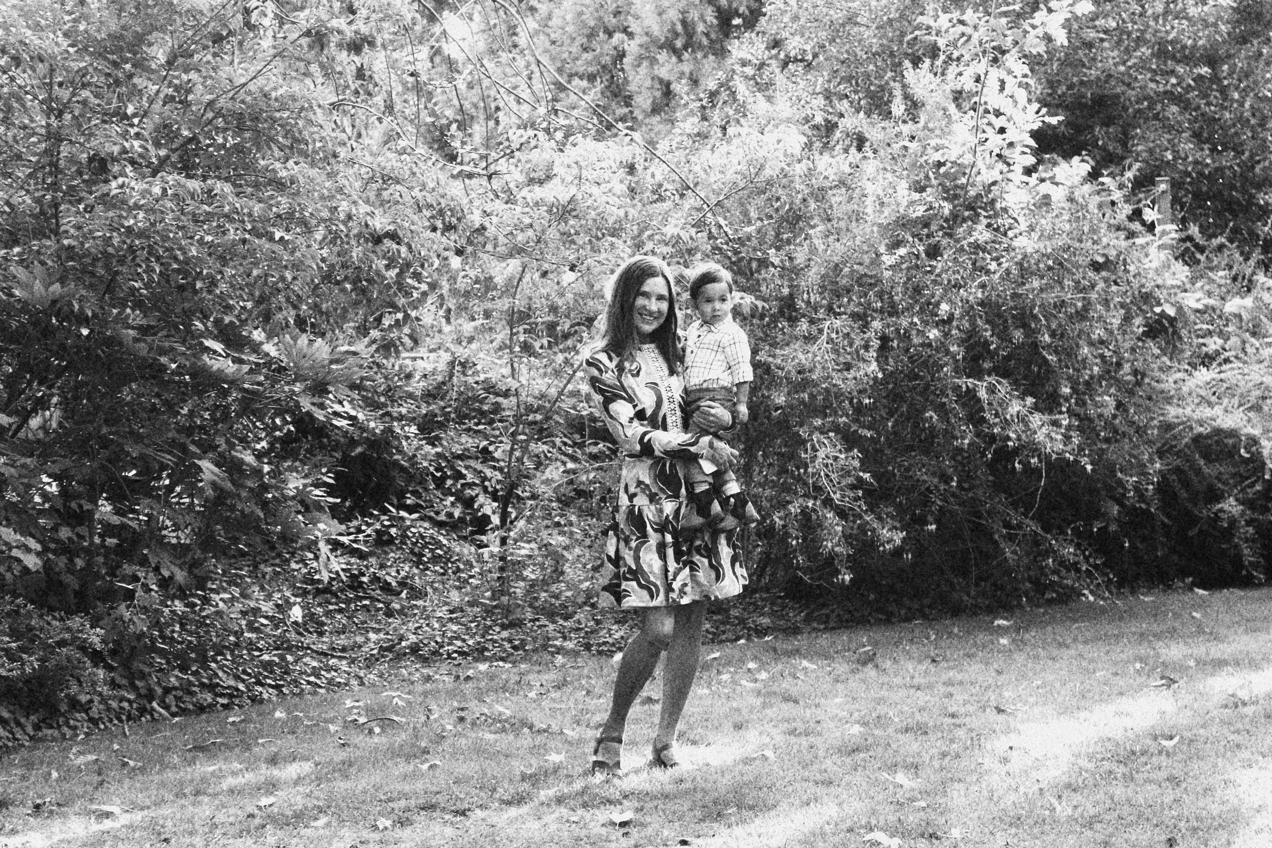 A woman holding a young boy standing on grass in front of dense bushes and trees.
