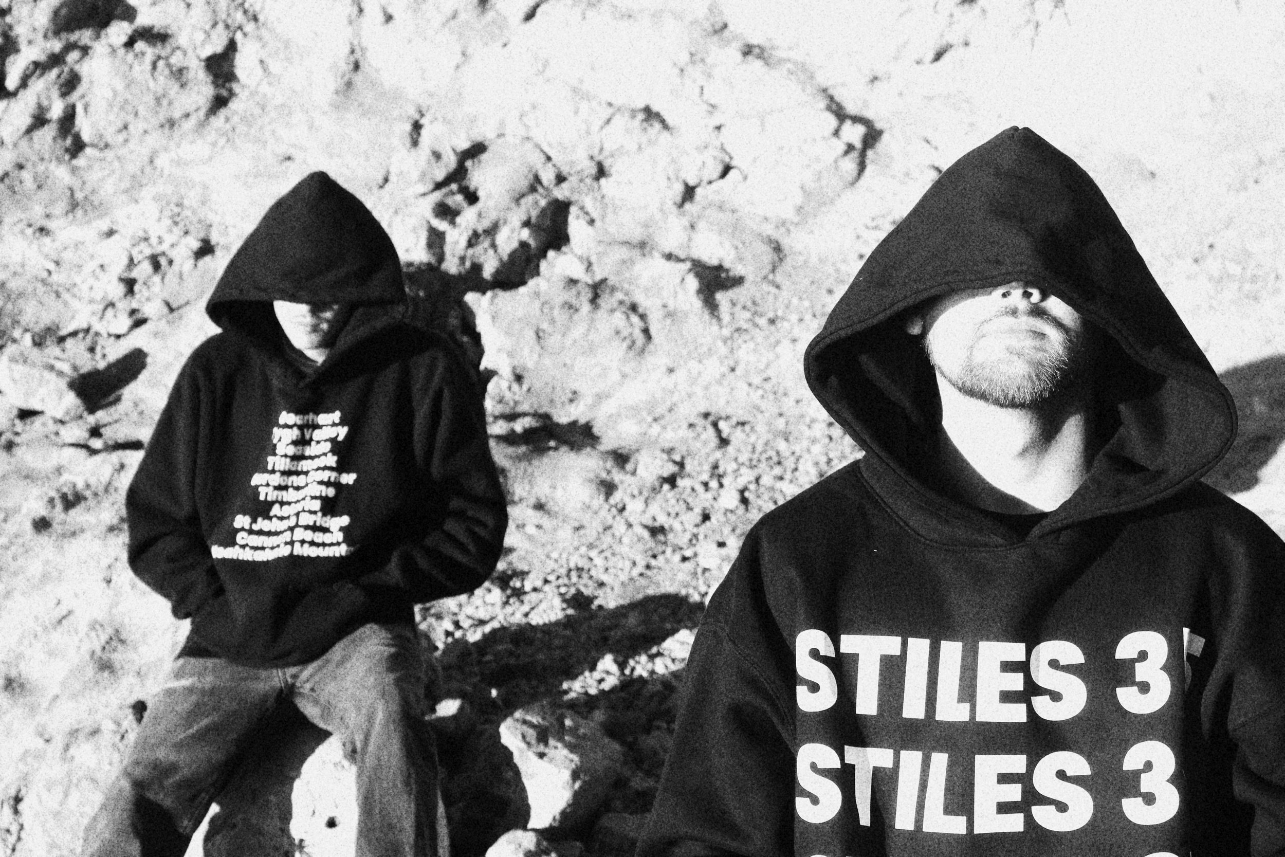 Two men with hooded sweatshirts, one sitting and one standing, on a rocky beach at night under a moonlit sky. The man in the foreground wears a hoodie with the text 'STILES 3' printed on it.