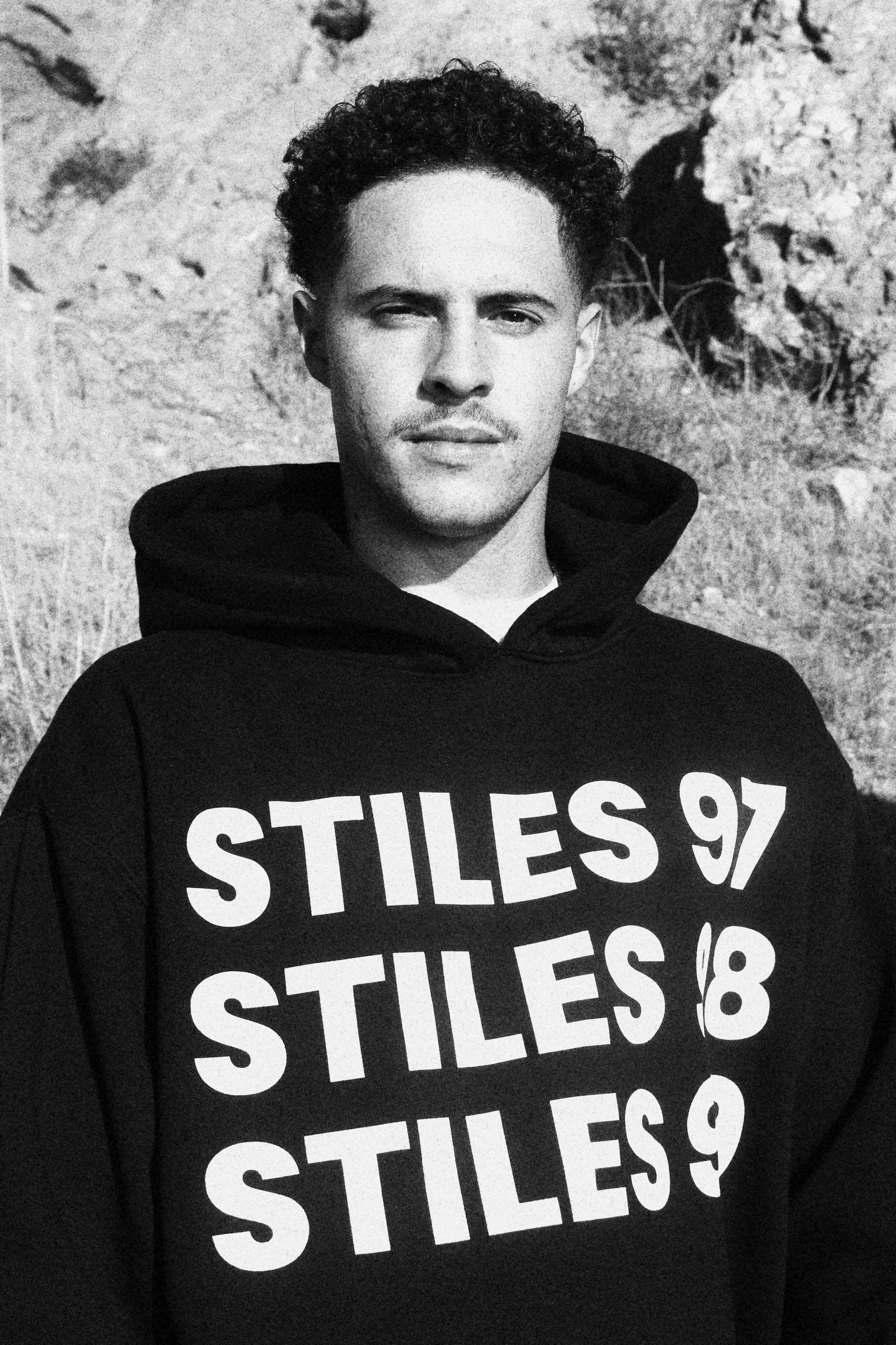 Black and white photo of a young man with curly hair wearing a hoodie, standing outdoors in front of a rocky landscape.