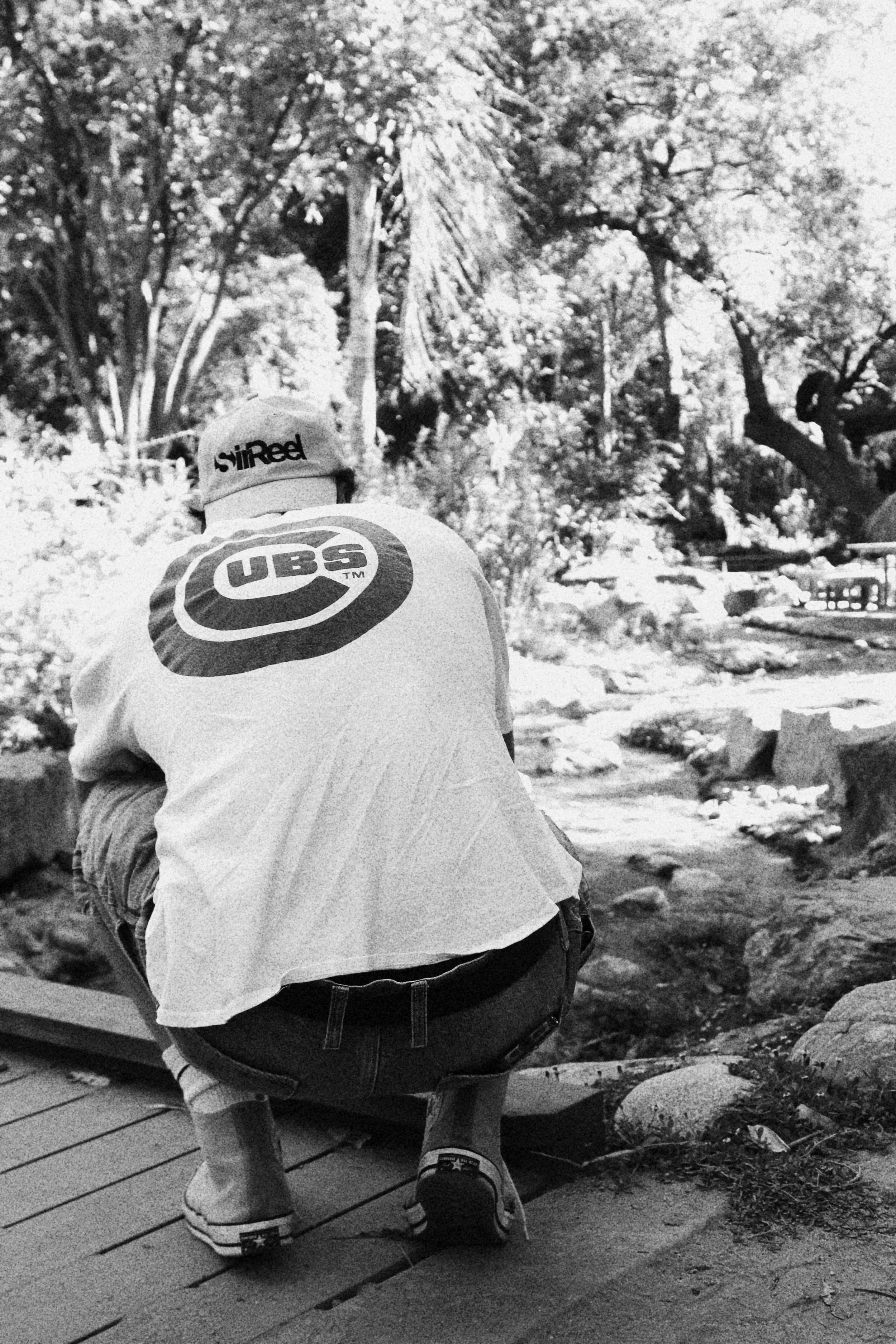 Person crouching near a small pond in a natural setting with trees and rocks in the background, wearing a cap and T-shirt with a logo.