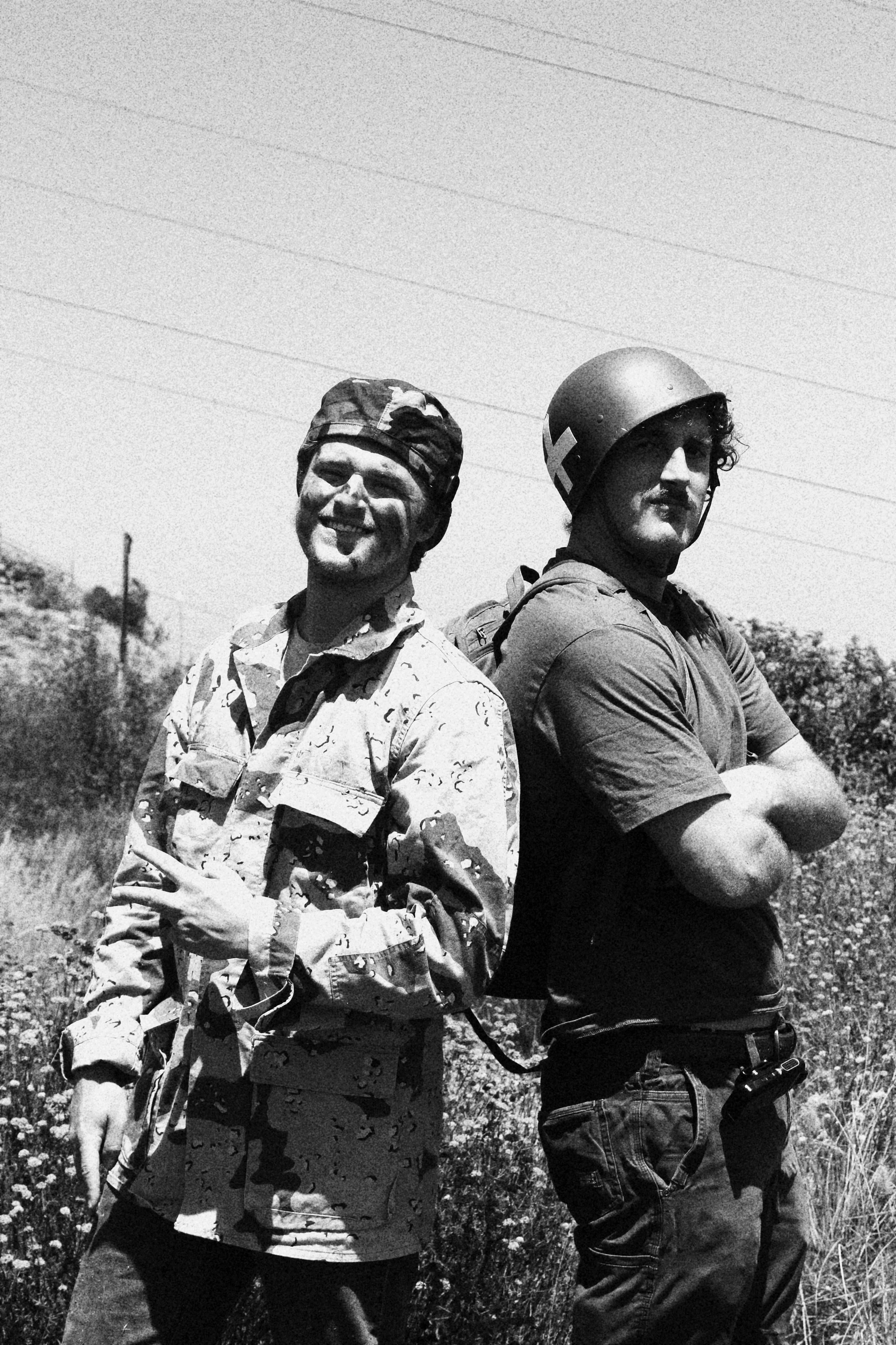 Two men stand back-to-back outdoors in a field, one in military camouflage with a smile and peace sign gesture, the other in a helmet and casual clothing with arms crossed.