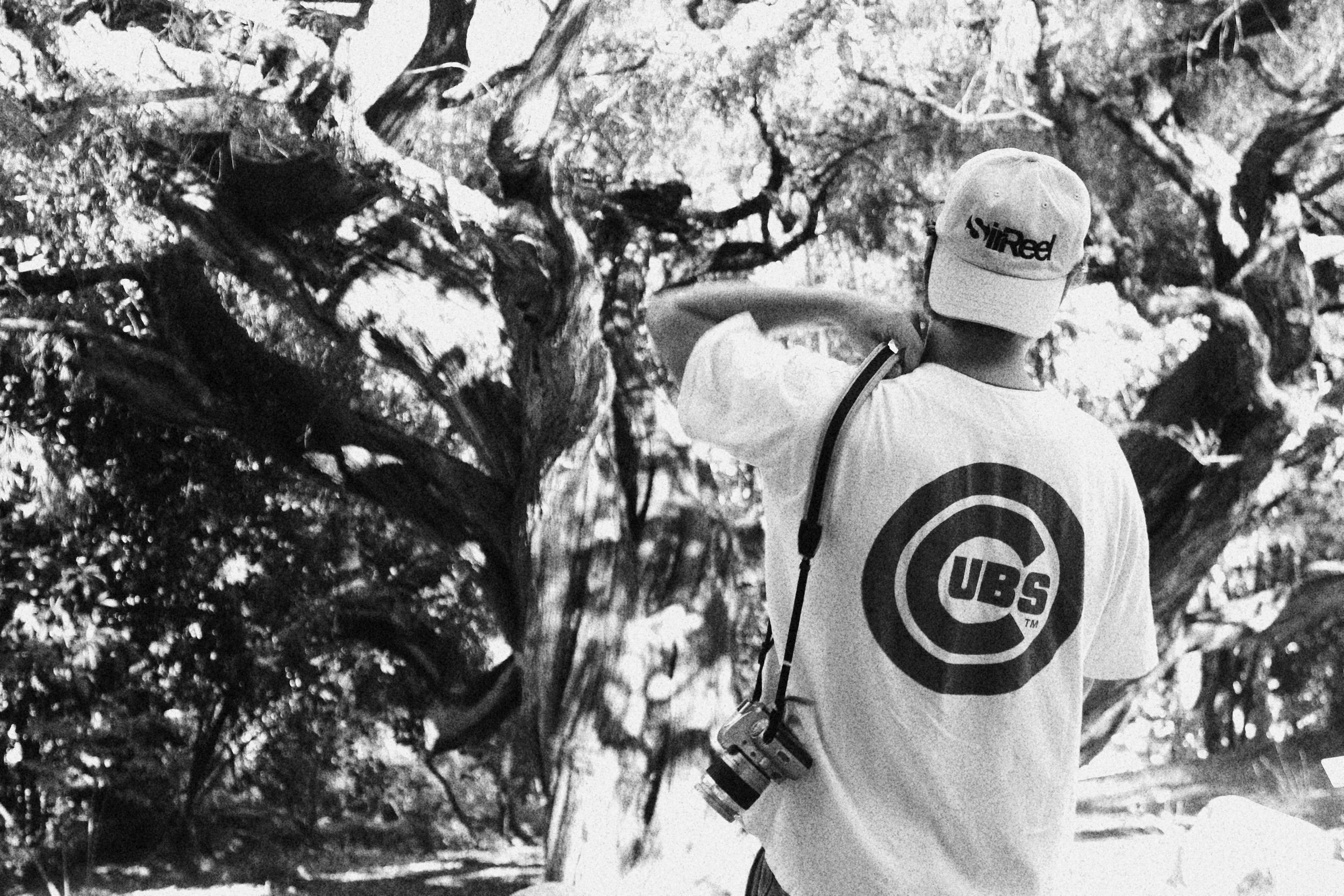 Person wearing a cap and t-shirt with the Chicago Cubs logo, standing outdoors near a large tree, holding a camera strap over their shoulder.