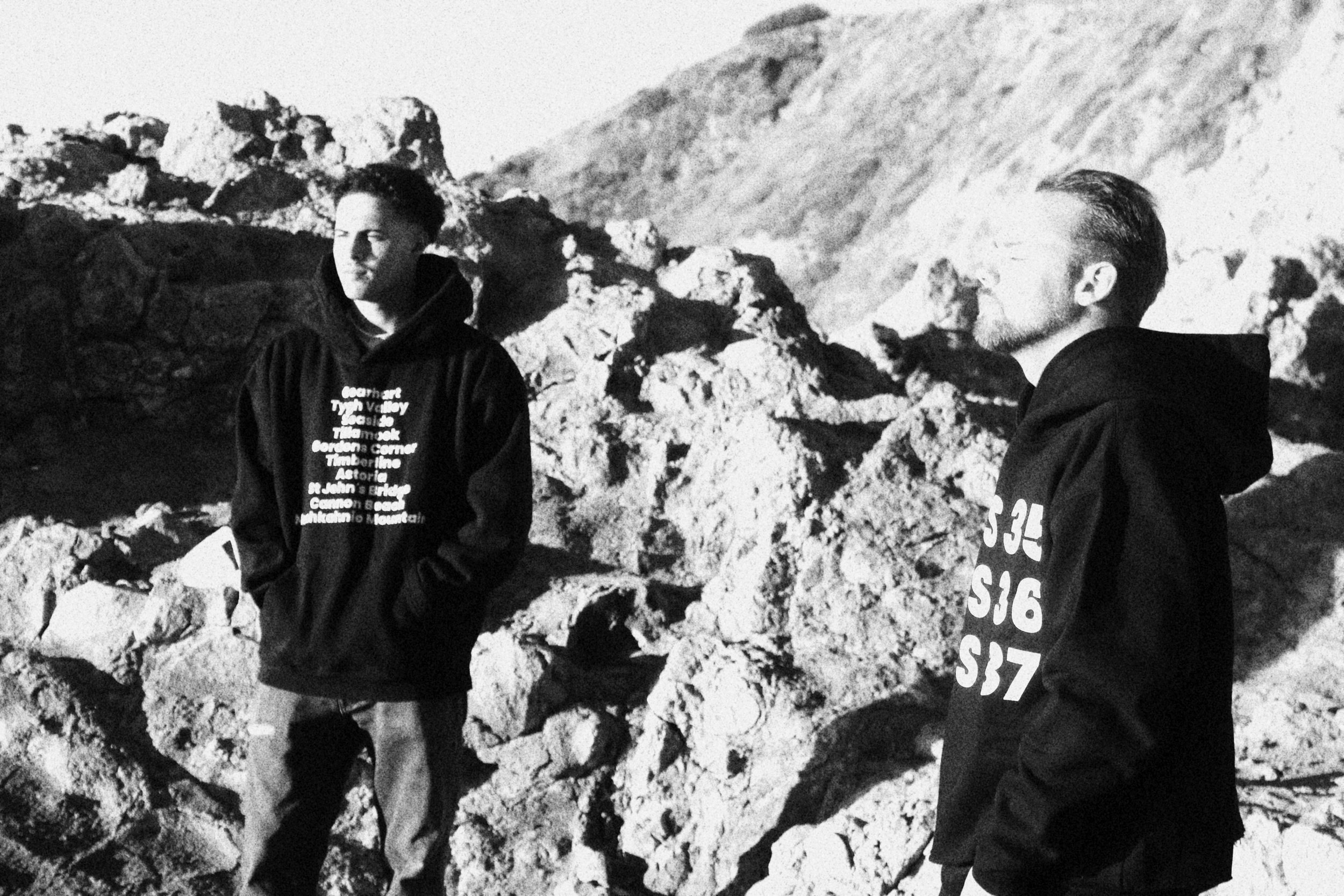 Two men standing outdoors among rocks in a mountainous area, wearing hoodies and looking away from the camera.