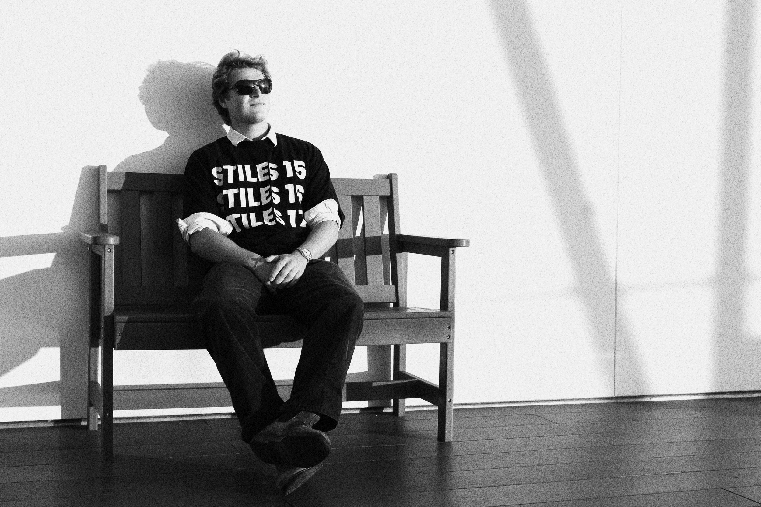 Black and white photo of a young man sitting on a wooden bench against a plain wall. He is wearing sunglasses, a T-shirt with the repeated word 'TILES 15', a collared shirt underneath, dark pants, and a watch. His hands are resting on his lap, and hi