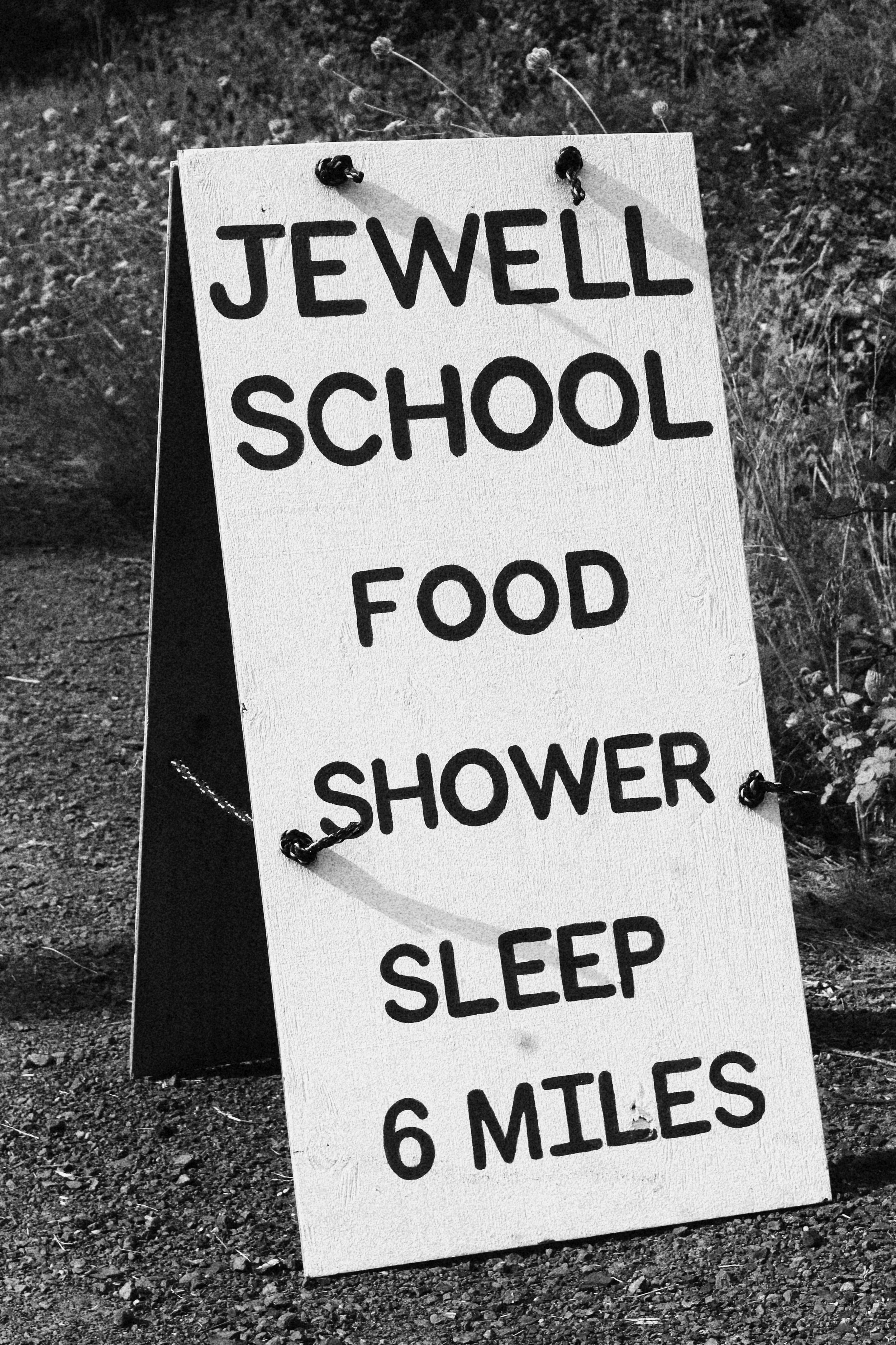 A white signboard on the ground with black handwritten text listing items: Jewel School, Food, Shower, Sleep, 6 miles, placed outdoors on dirt with grass and plants in the background.