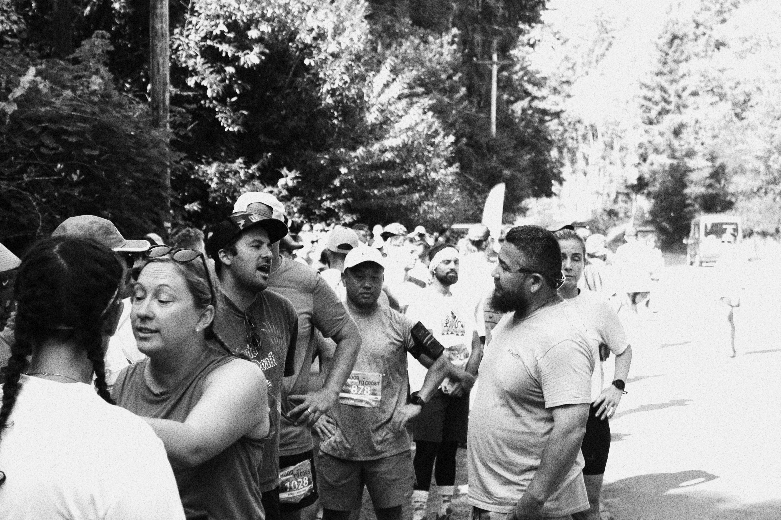 A group of runners gathered outdoors, some engaging in conversation, with trees in the background, indicating a race or marathon event.