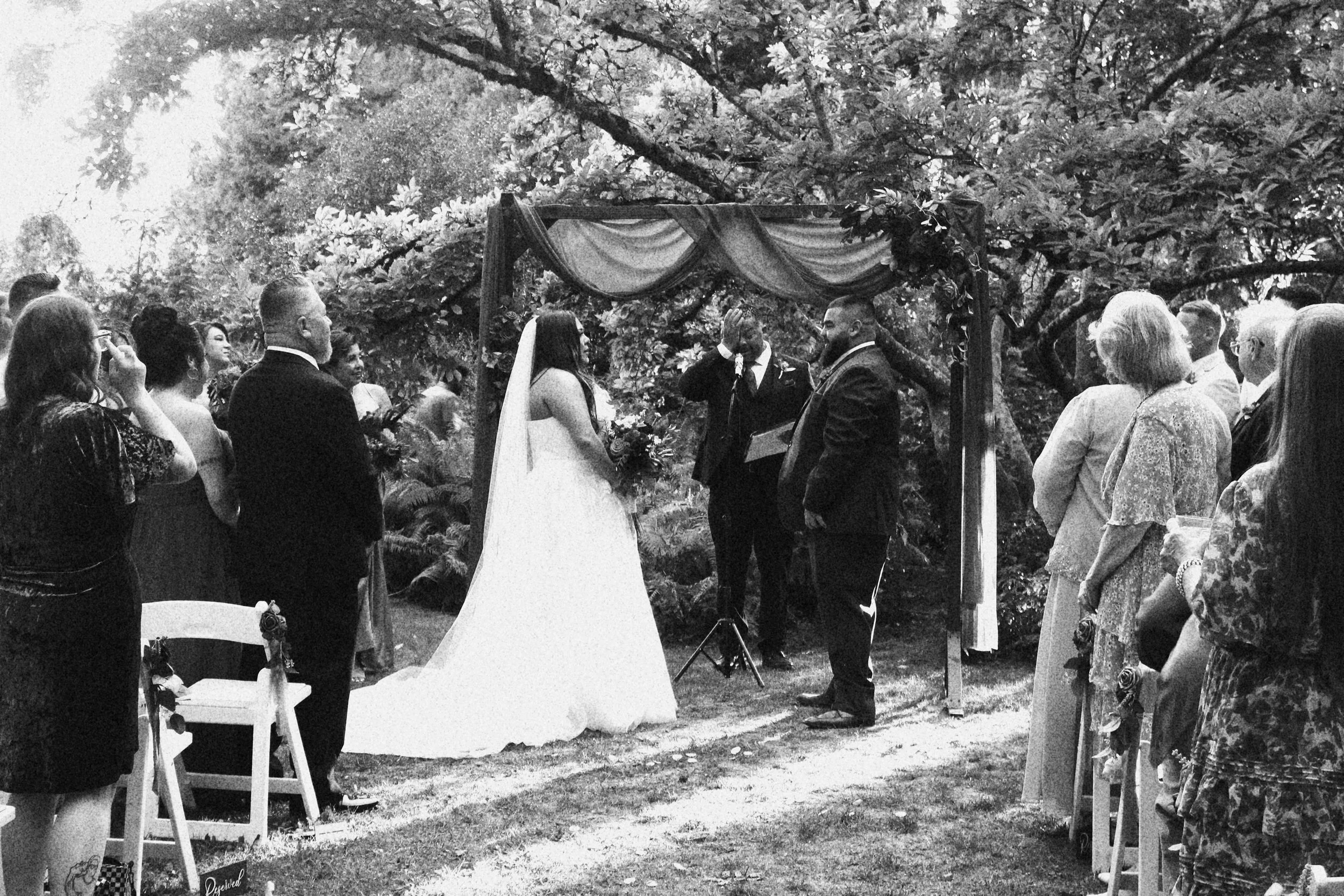 A black and white photo of a wedding ceremony taking place outdoors, with the bride and groom standing under an arch decorated with drapery and flowers, surrounded by guests.