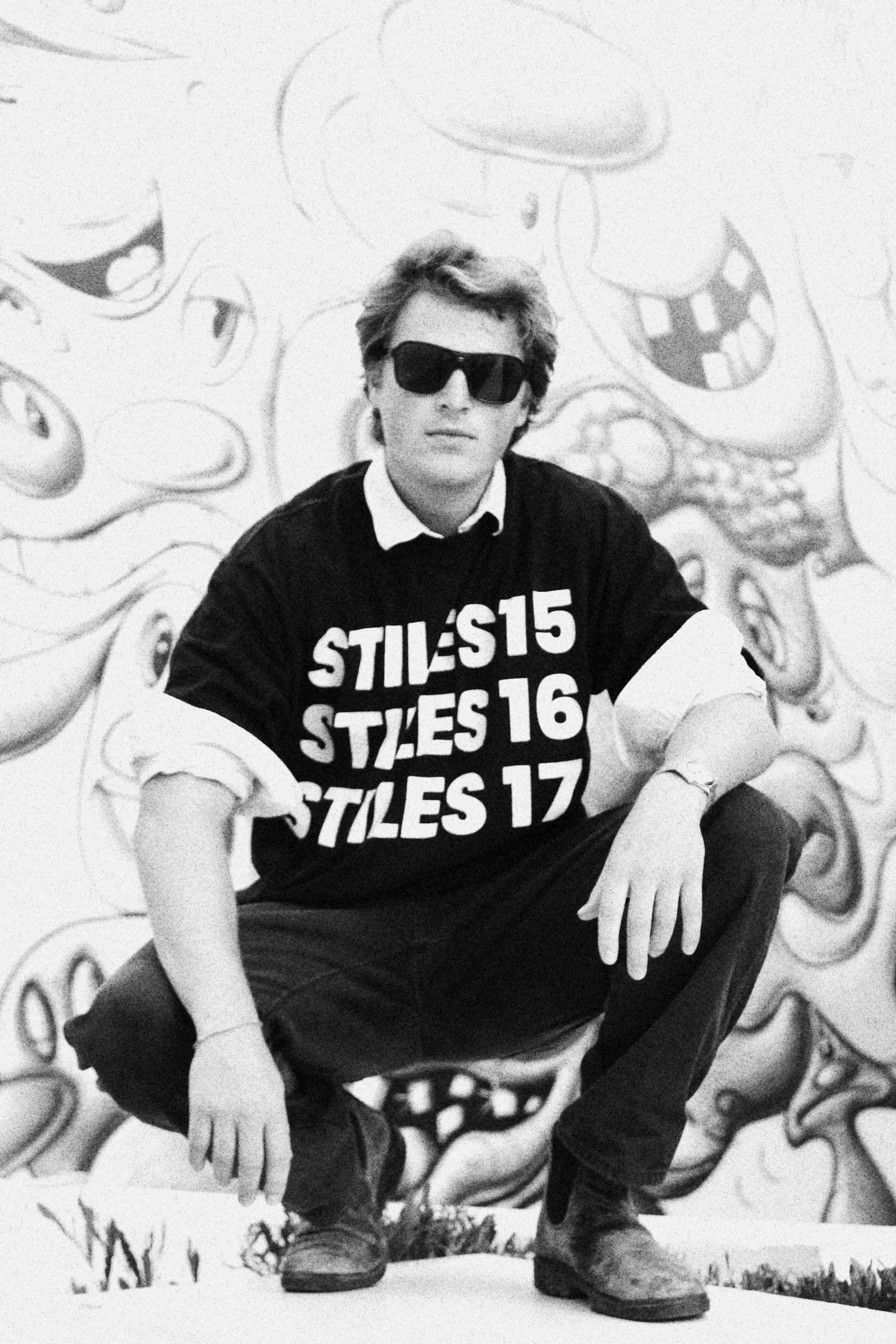 A black-and-white photo of a young man wearing sunglasses, a layered shirt, and a T-shirt with the text 'STILES 15, STILES 16, STILES 17.' He is crouched on the ground in front of a graffiti-style mural with cartoon faces.