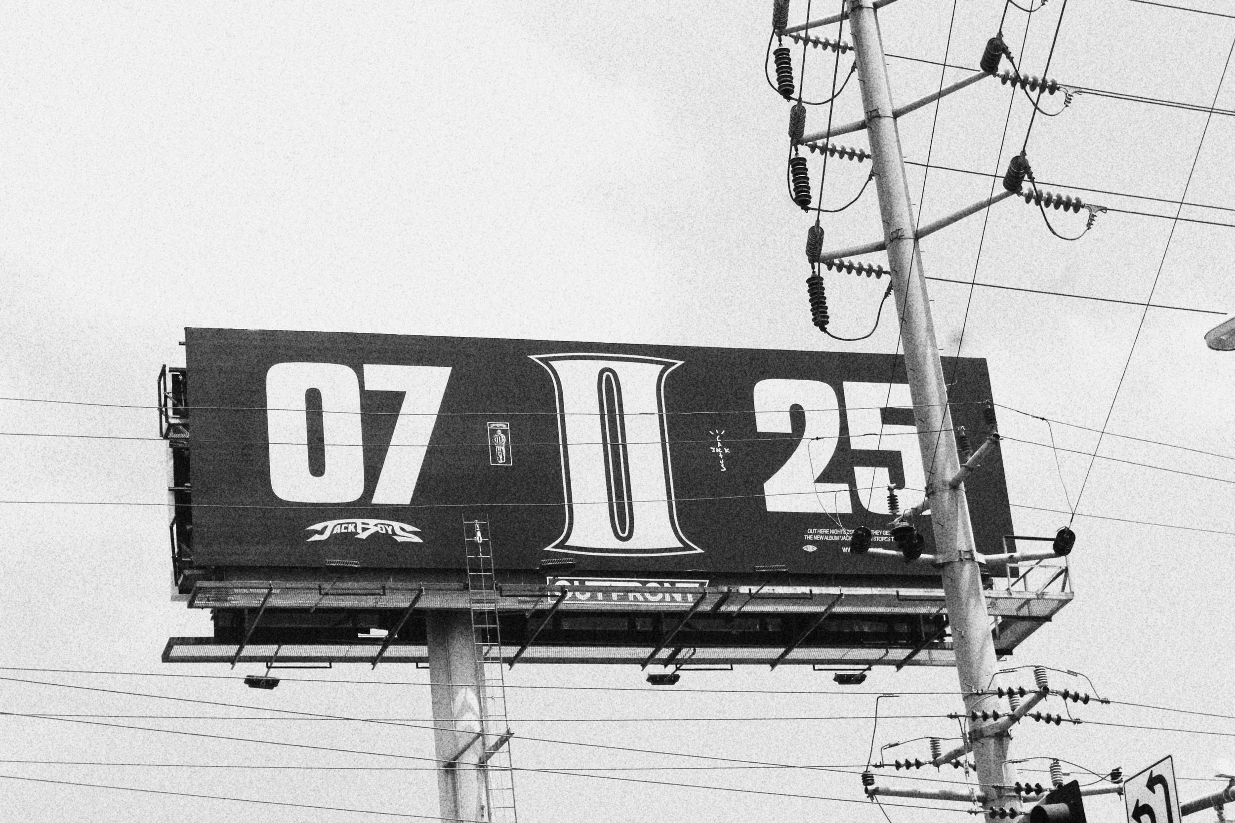 Black and white billboard displaying the numbers 07, 10, and 25 with a large letter I in the center, mounted on a pole alongside utility power lines.